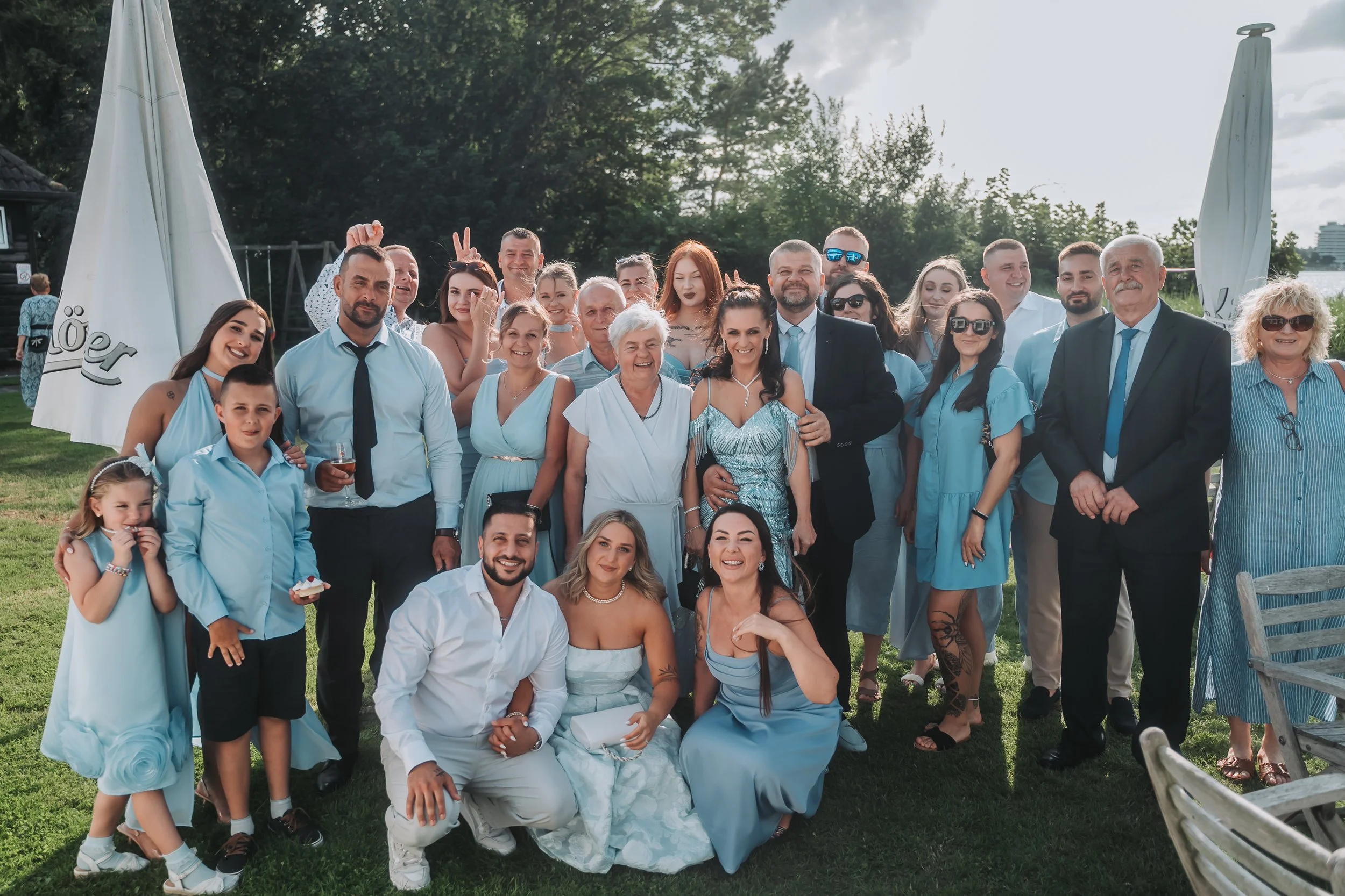 Group of people at an outdoor event, smiling and posing for a photo, dressed in light blue and white, with trees and umbrellas in the background.