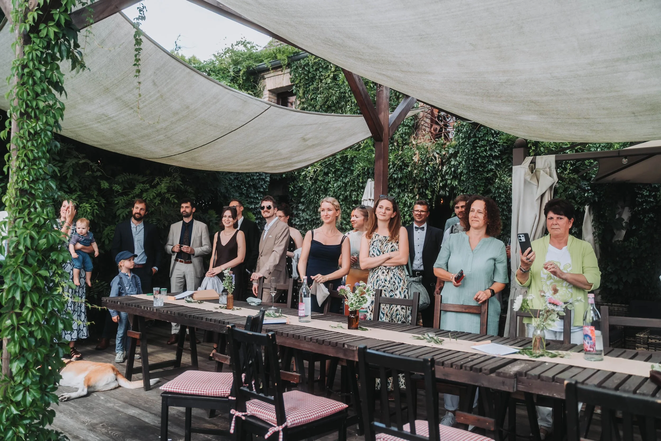 A group of people gathered outdoors under a canopy for a social event or celebration, with tables and chairs, surrounded by greenery.