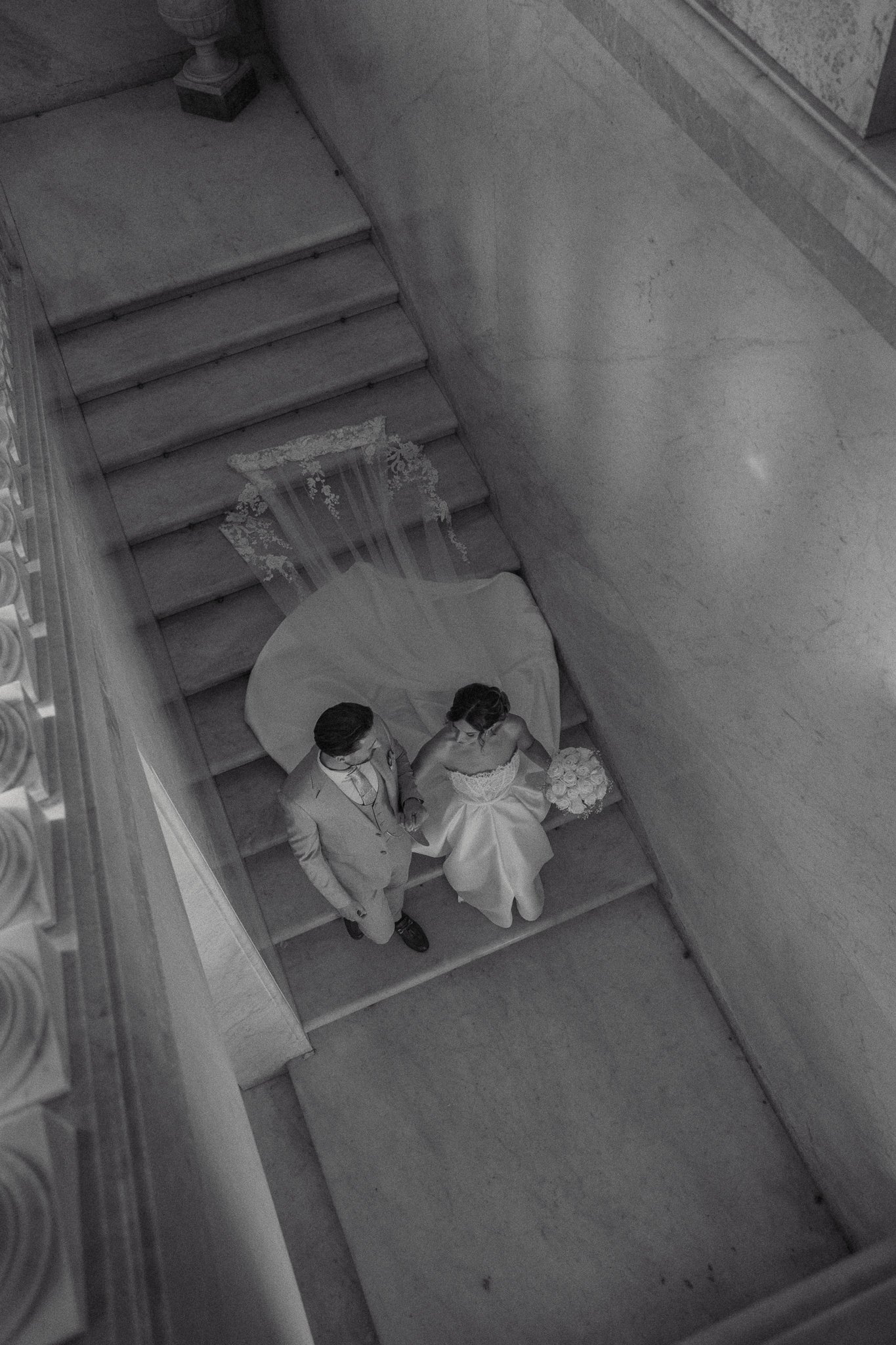A bride and groom holding hands on a staircase, with the bride holding a bouquet, dressed in wedding attire, and the groom in a suit, seen from above in black and white.