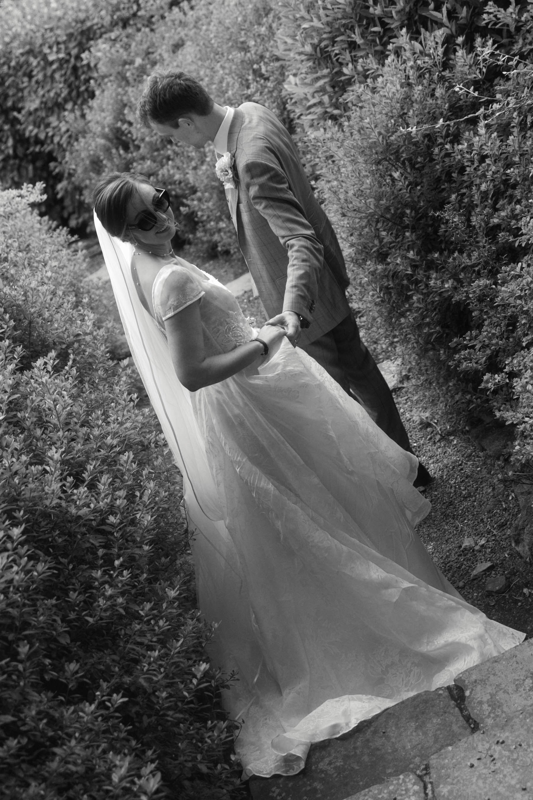 A bride and groom holding hands and smiling in a garden path with bushes on both sides. Black and white photo.