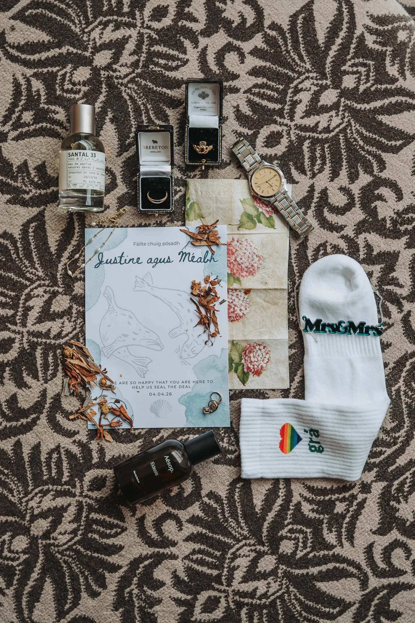 Flat lay of personal items on a patterned carpet, including perfume, jewelry, a wristwatch, a sock with rainbow heart and healing text, a small box with jewelry, herbal plants, a greeting card, and a bottle of skincare product.