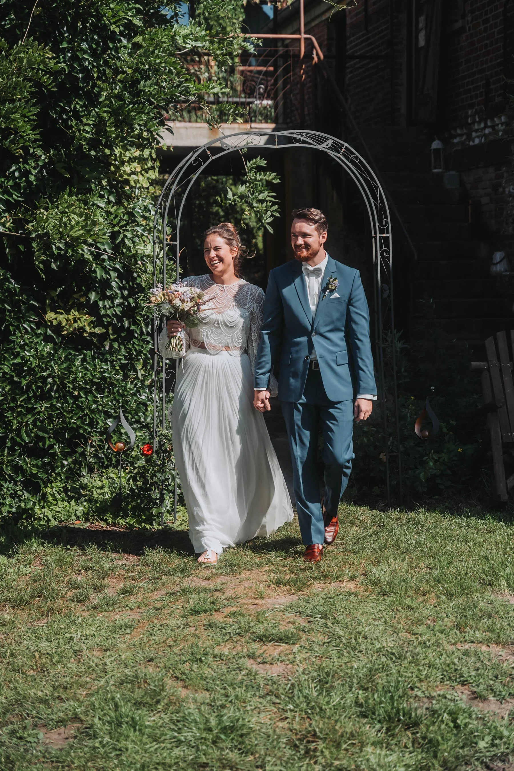 A bride and groom walking hand in hand outdoors. The bride is wearing a white wedding dress and holding a bouquet, smiling. The groom is dressed in a blue suit with a white shirt, also smiling. They are walking through a garden archway, surrounded by