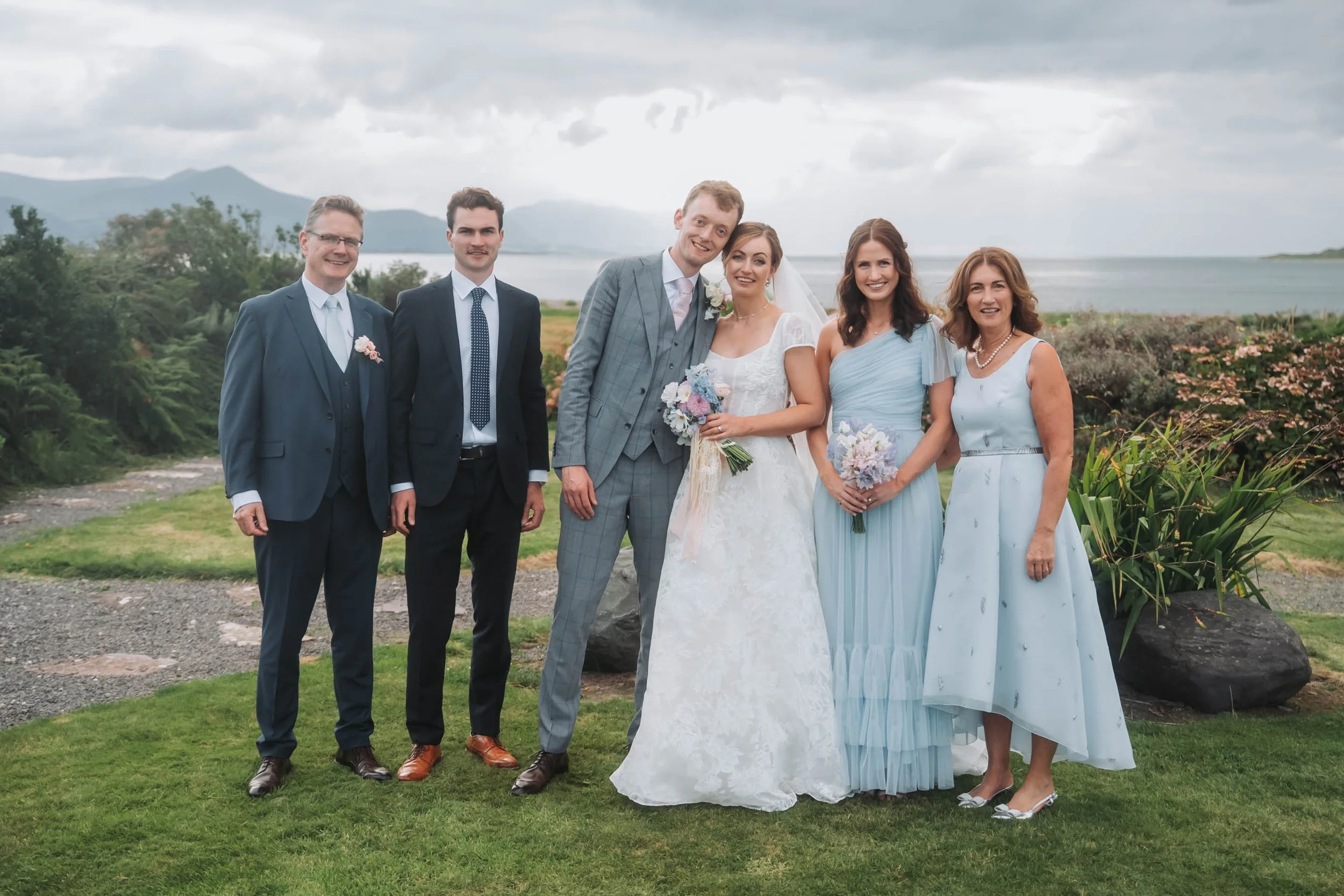 A group of six people at a wedding outdoors, including the bride and groom in the center, with three women and two men, standing on grass near a body of water, smiling.