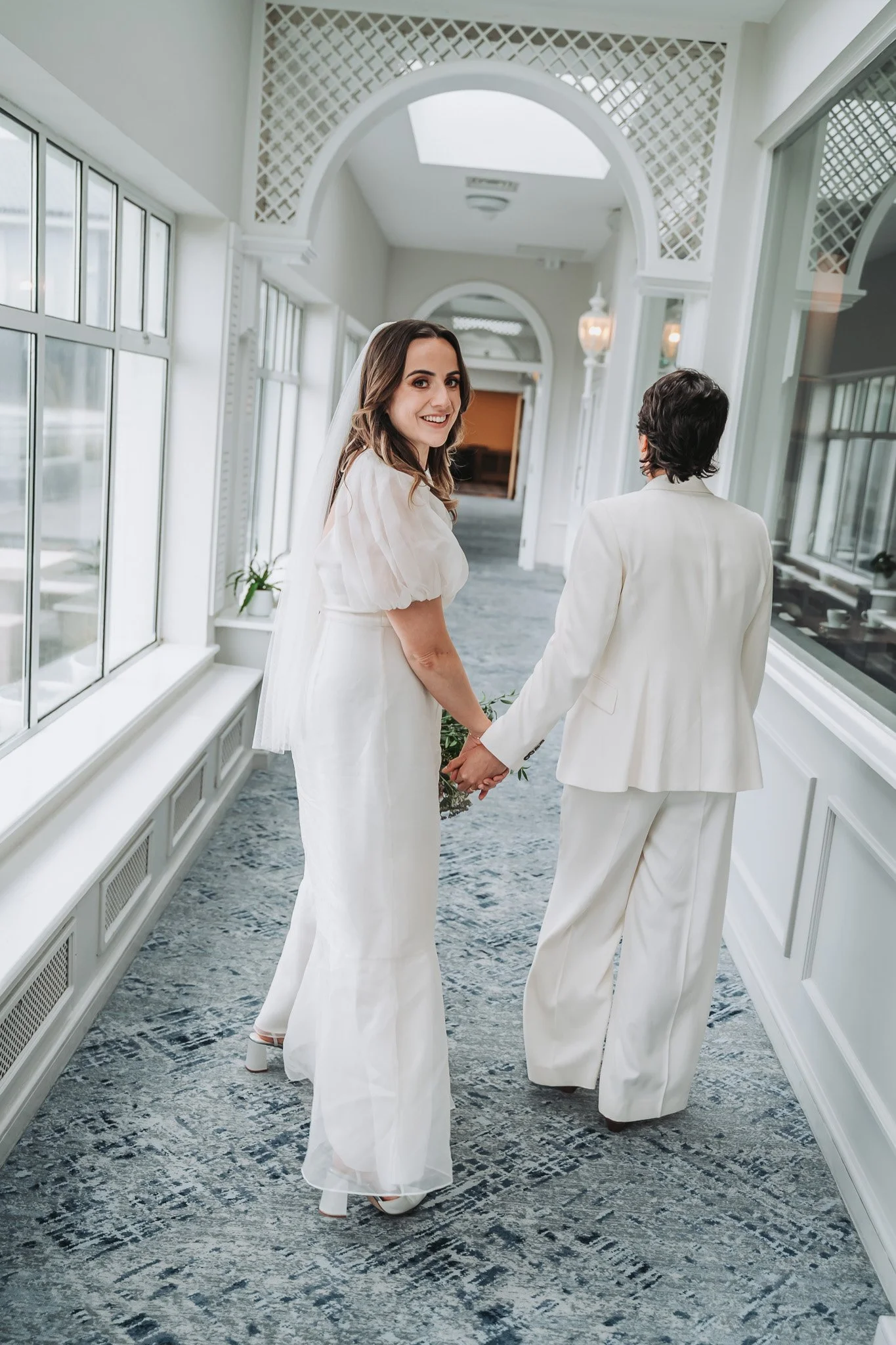 A bride with brown hair and a white dress holding hands with a person in a white suit inside a bright, elegant corridor with large windows and arched doorways.