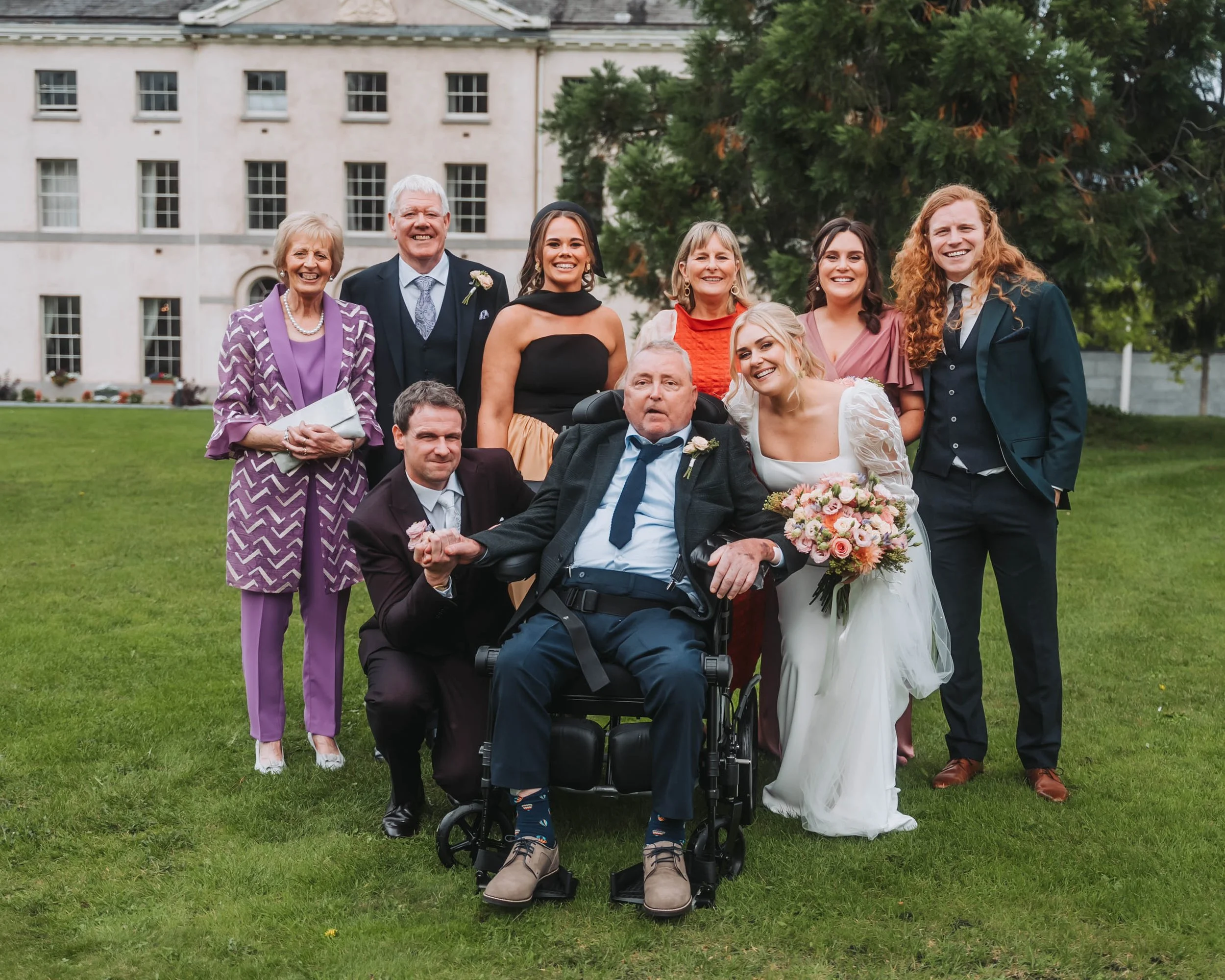 A group of smiling family members and friends gathered outdoors on a lawn near a large building, celebrating a wedding. The bride, dressed in a white gown, holds a bouquet of flowers, and is smiling. The groom, in a dark suit and tie, is seated in a 