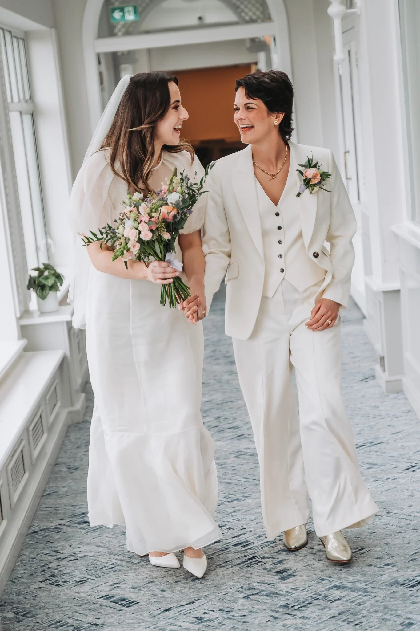 Two women walking together, smiling, in an indoor setting with white walls and windows. One is wearing a white wedding dress and holding a bouquet of flowers, the other is dressed in a white pantsuit with floral boutonnière.