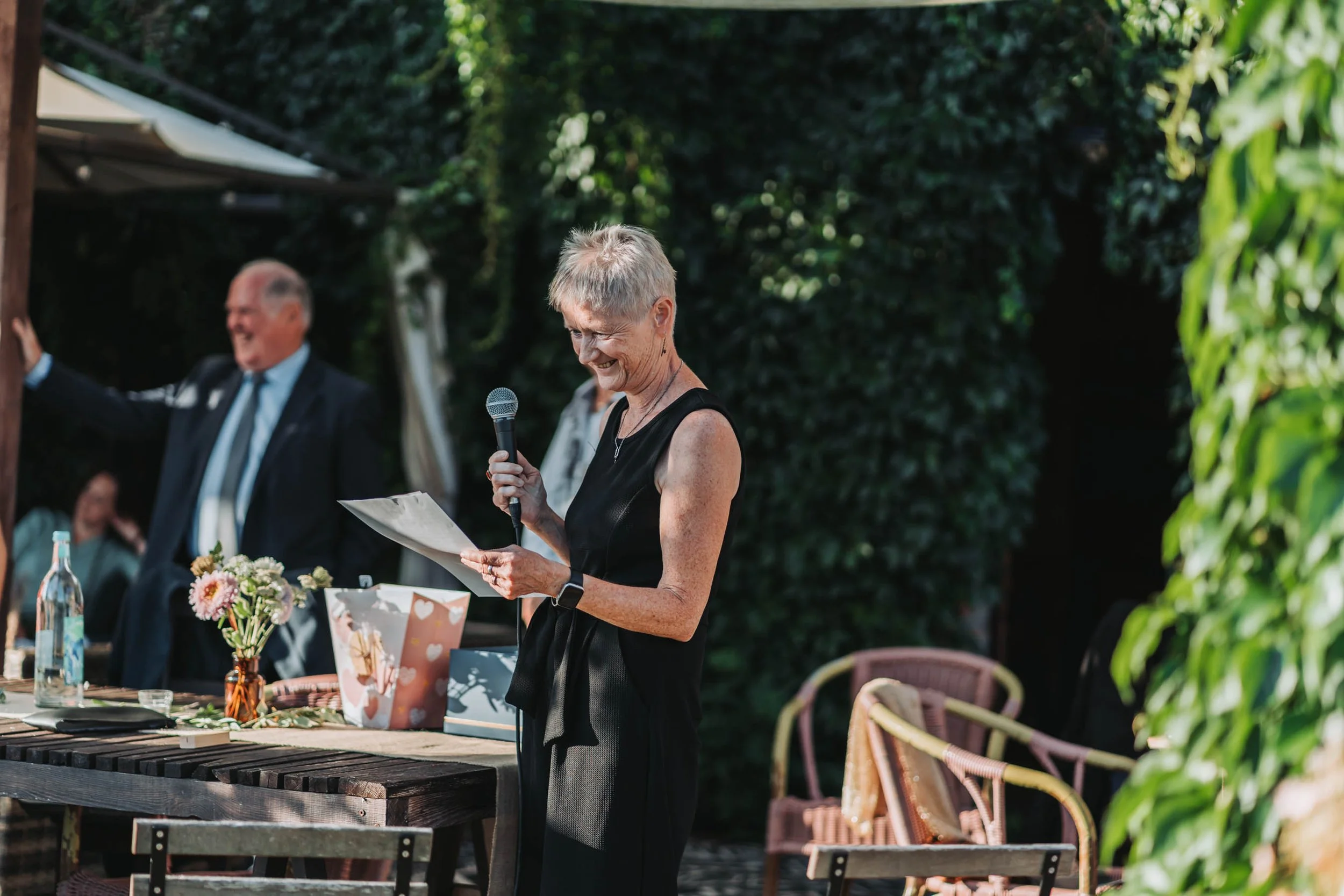 A woman with short gray hair, wearing a black sleeveless dress, is holding a microphone and reading from a sheet of paper at an outdoor event. There are two older men in suits in the background, and a table decorated with flowers and gift bags.