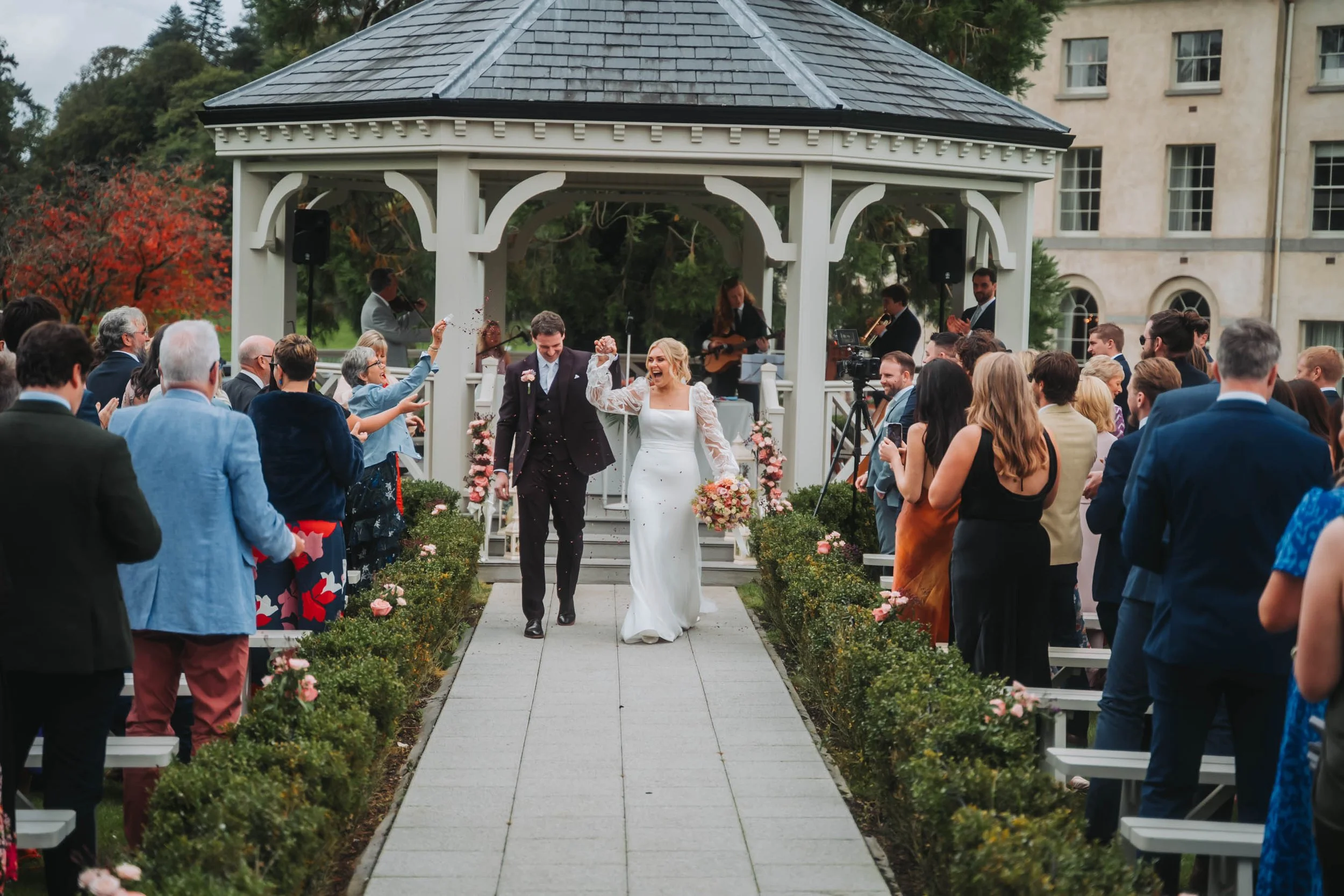 A wedding celebration with a bride and groom walking down an outdoor aisle surrounded by guests. The bride is laughing and holding a bouquet, and the groom is smiling. There is a band playing under a gazebo in the background, and the scene is decorat