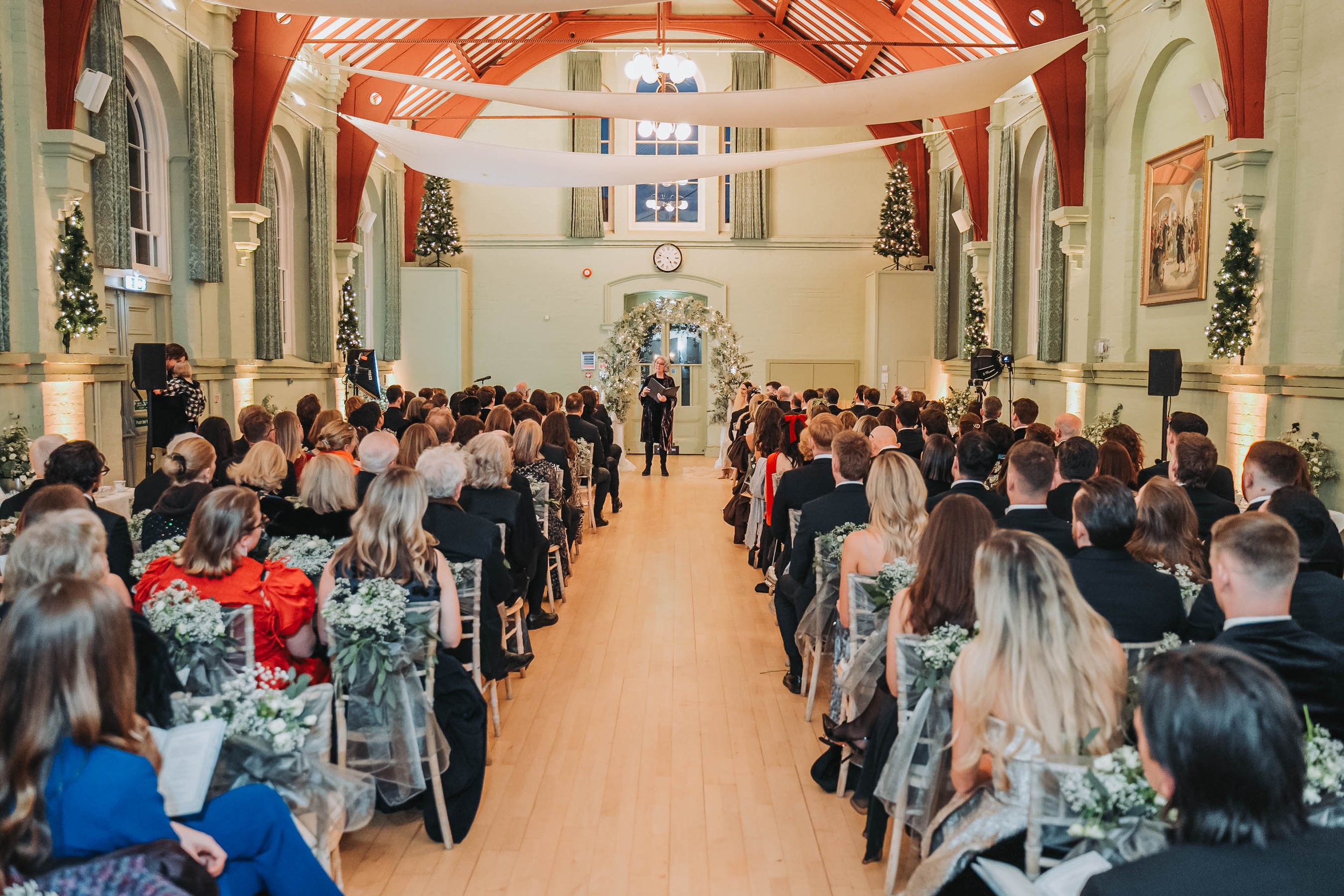 A large indoor gathering with seated attendees watching a speaker at a podium decorated with Christmas trees and lights, in a room with high ceilings, arched windows, and festive decorations.