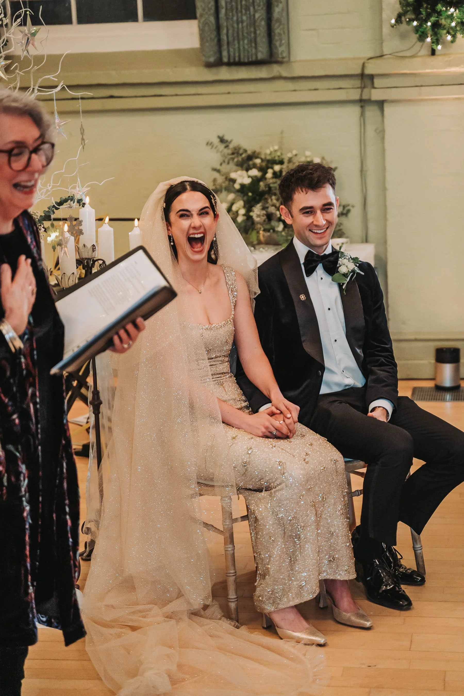 A bride and groom sitting together during their wedding ceremony, both smiling, with a woman reading vows or giving a speech nearby. The bride is in a glittery gold wedding dress with a matching veil, and the groom is in a black tuxedo with a bow tie