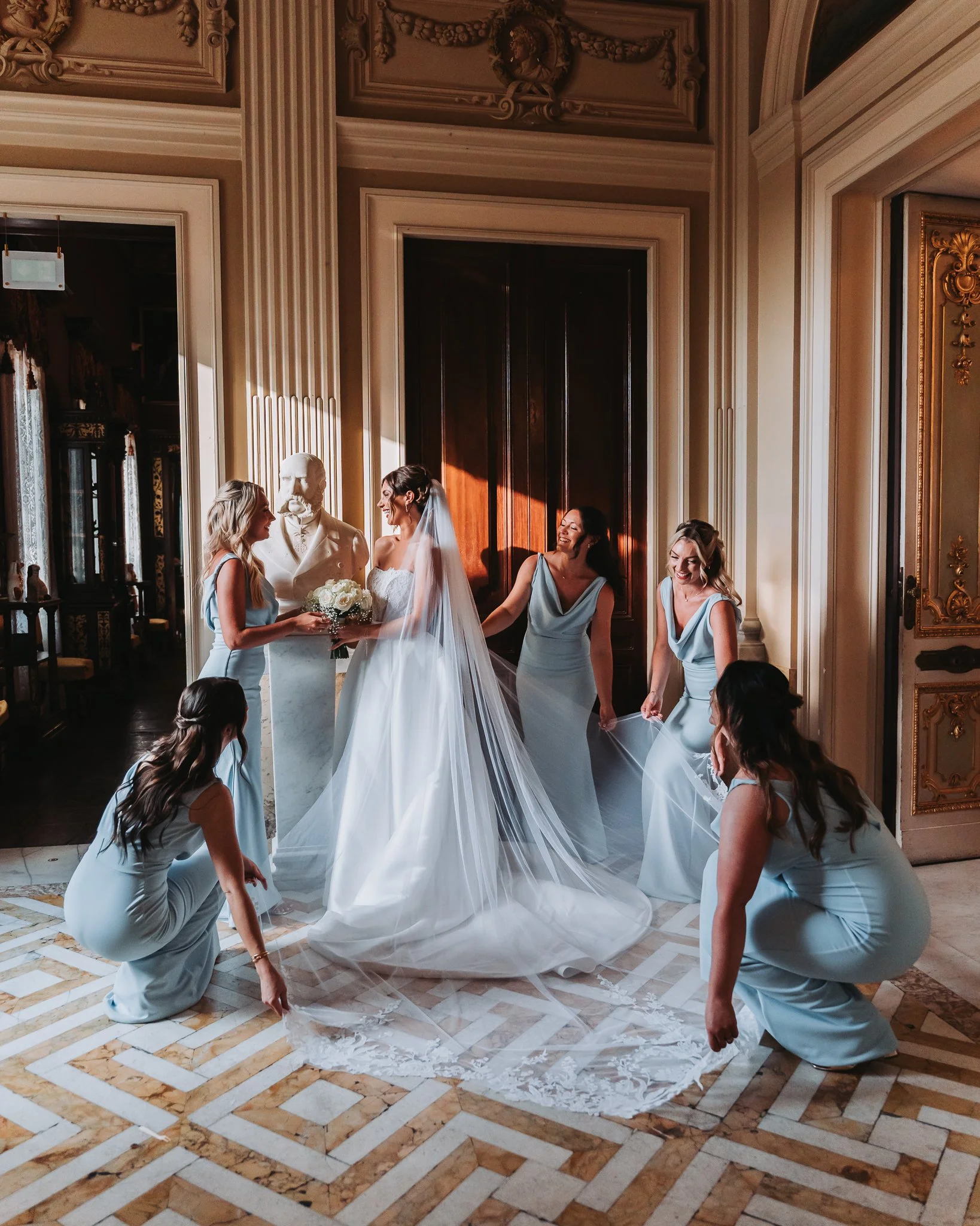 Bride and bridesmaids in a luxurious room with ornate moldings and marble floors. The bride, wearing a white wedding dress, is surrounded by bridesmaids in matching pale blue dresses, decorating her dress with the train and veil.