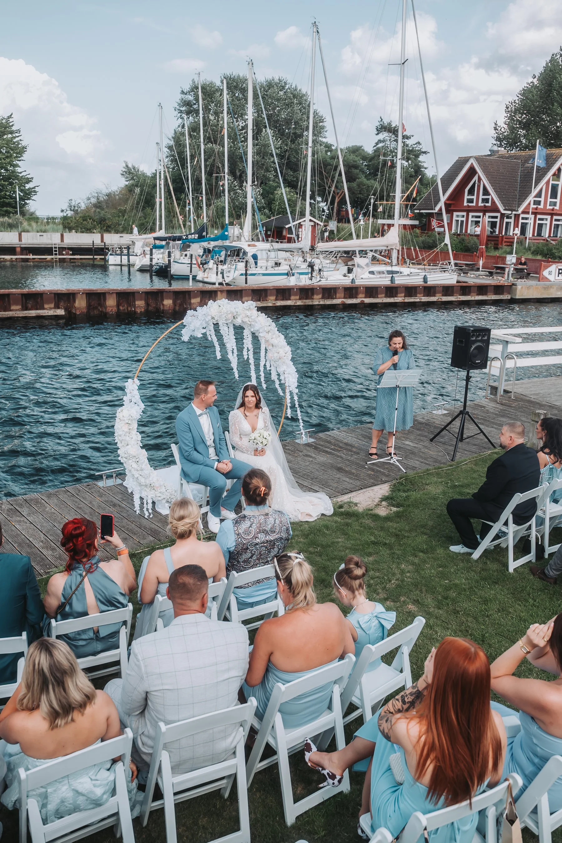 A wedding ceremony takes place outdoors on a wooden dock by a lake, with boats and a house in the background. The bride and groom are seated under a circular floral arch, and a woman is giving a speech at a microphone nearby. Guests are seated on whi