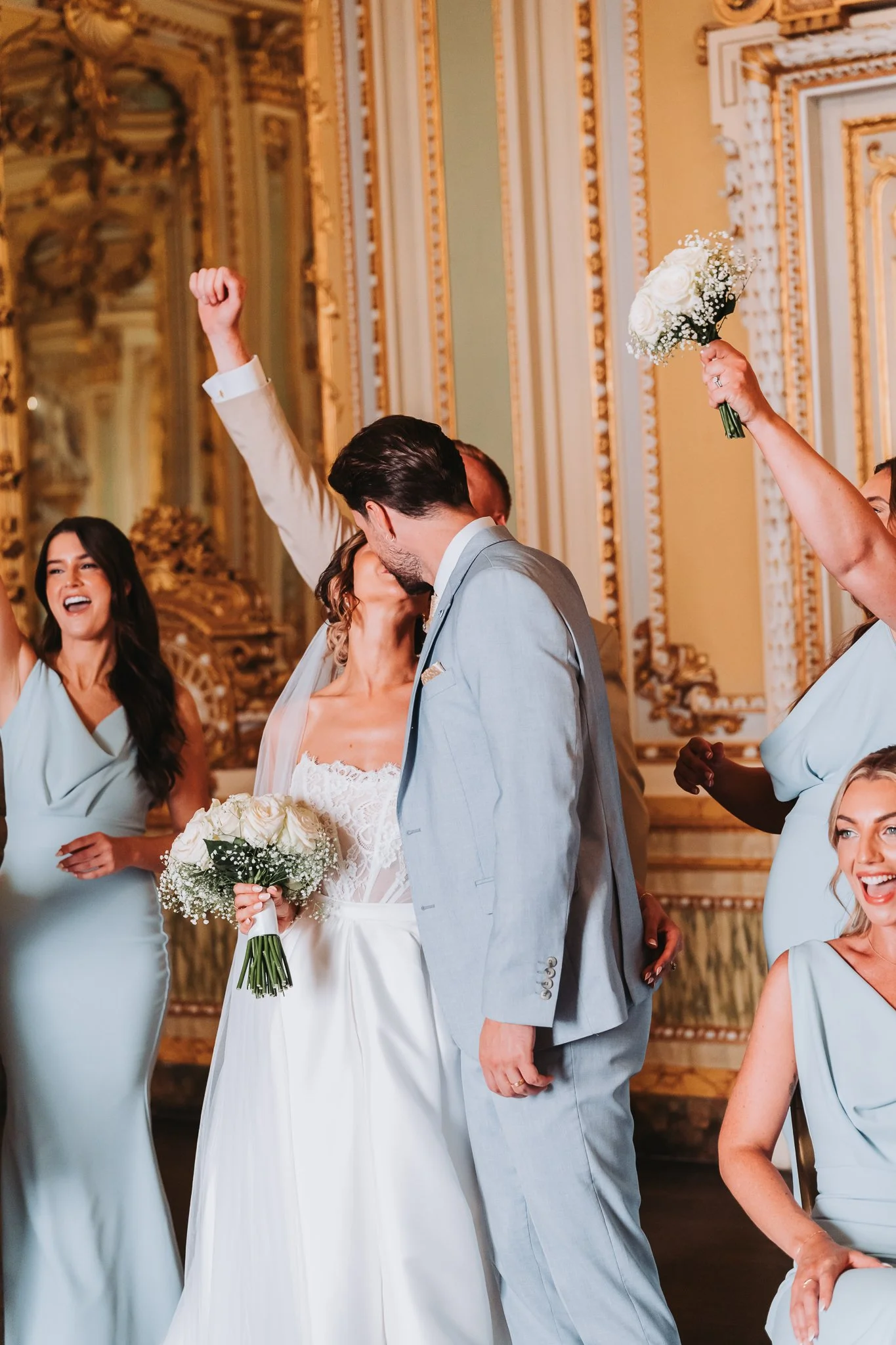 Wedding scene with a bride and groom kissing, surrounded by bridesmaids celebrating, one holding a bouquet of white roses, in an ornate ballroom with gold detailing.