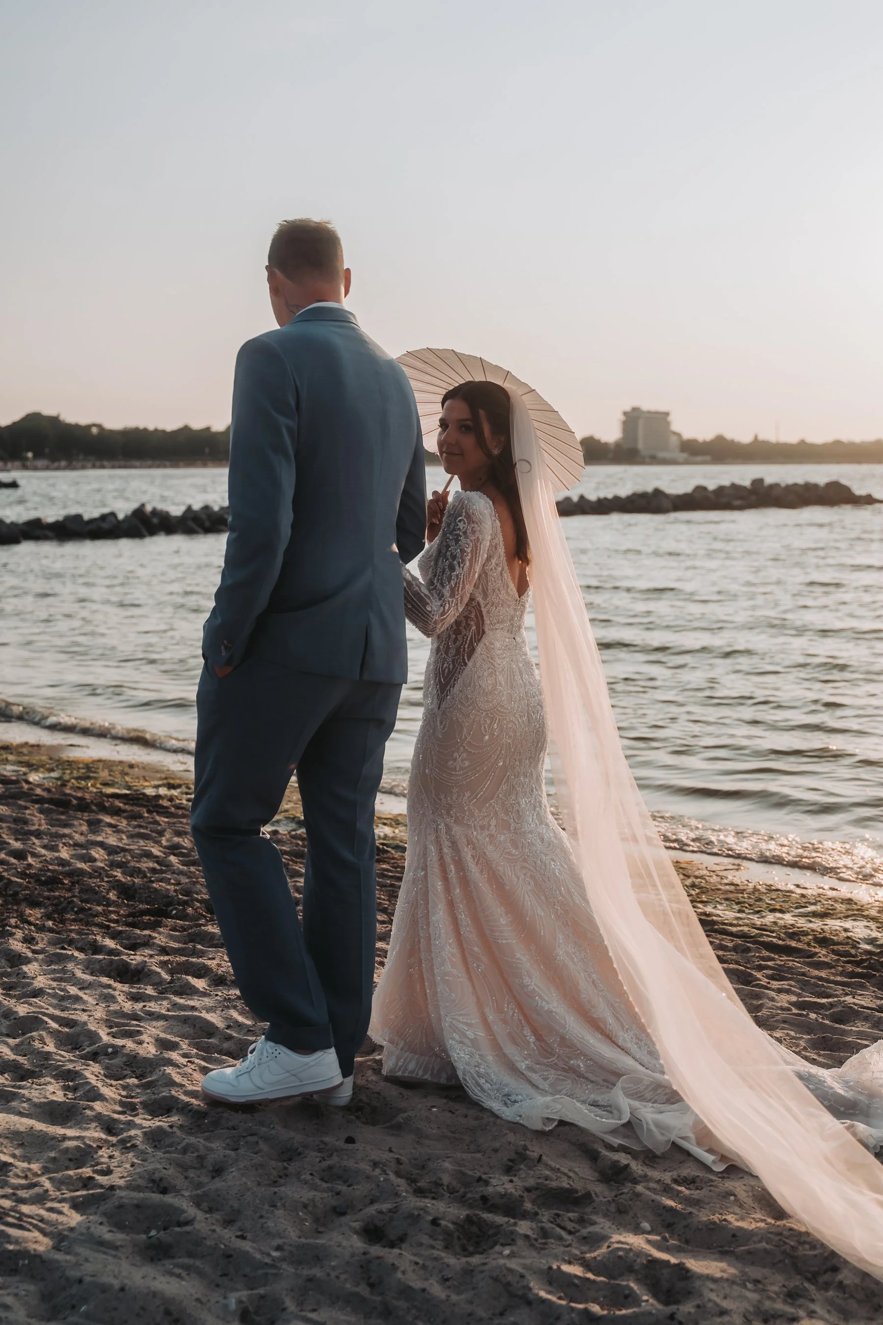 A bride in a white wedding dress holding a parasol and a groom in a blue suit standing on a beach near water during sunset.