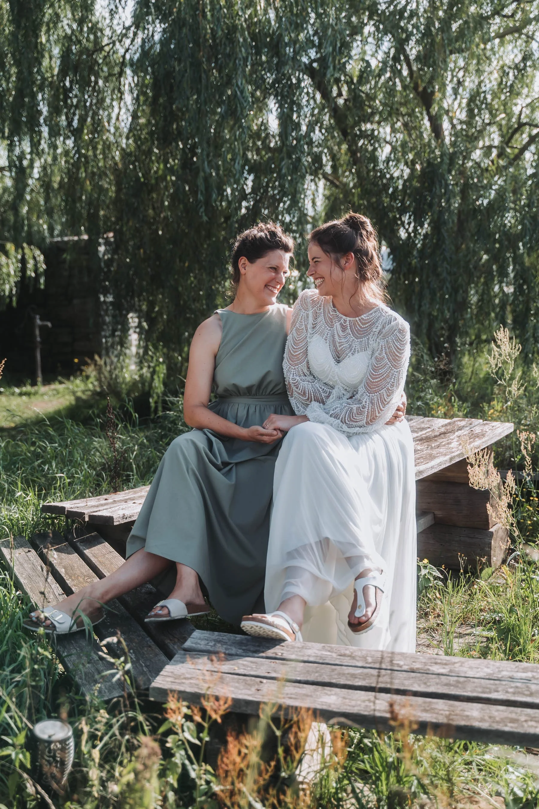 Two women sitting closely on a rustic wooden bridge outdoors, smiling and looking at each other, surrounded by greenery and trees.