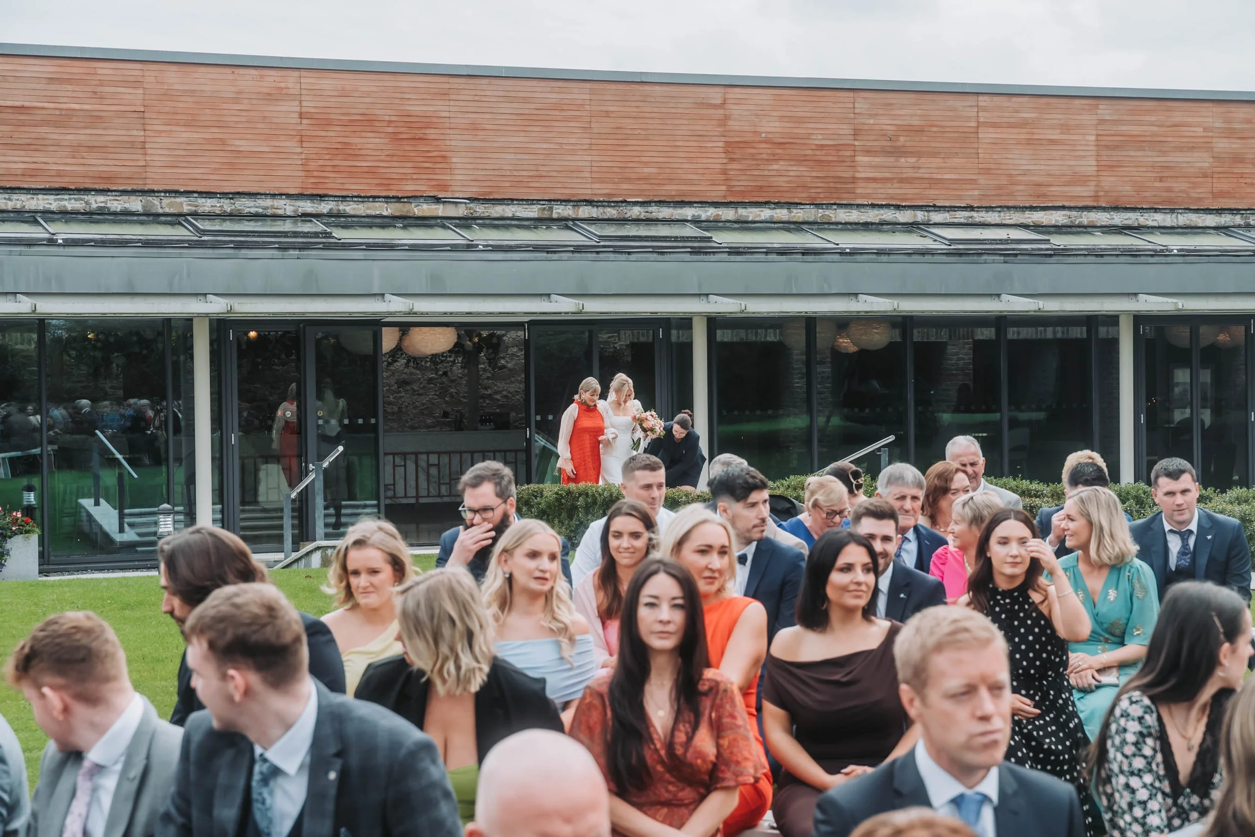 A large group of people dressed in formal attire seated outdoors for a wedding ceremony, with a couple standing on a platform in the background.