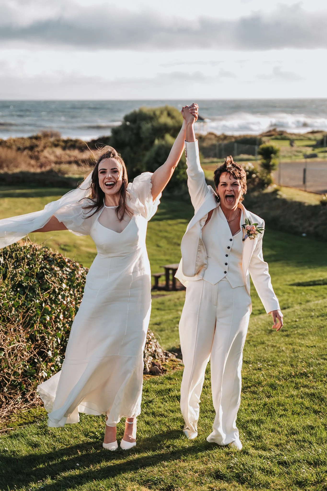Two women in white attire celebrating outdoors near the ocean, holding hands up in victory, with green grass, bushes, and ocean waves in the background.