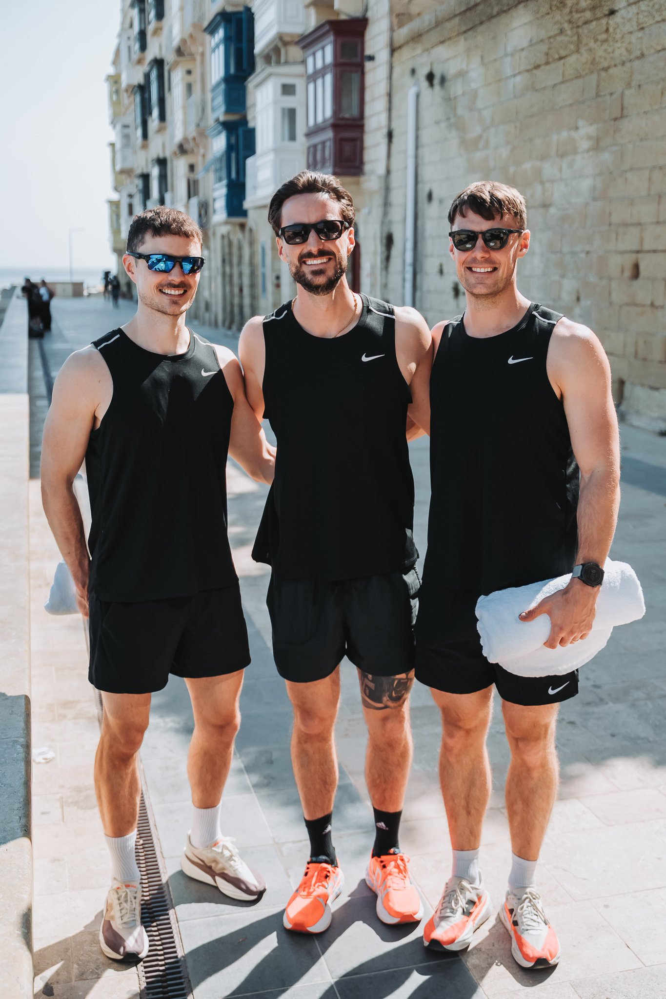 Three men in black athletic shirts and shorts standing outdoors near a brick building, smiling, wearing sunglasses and athletic shoes, with the ocean visible in the background.