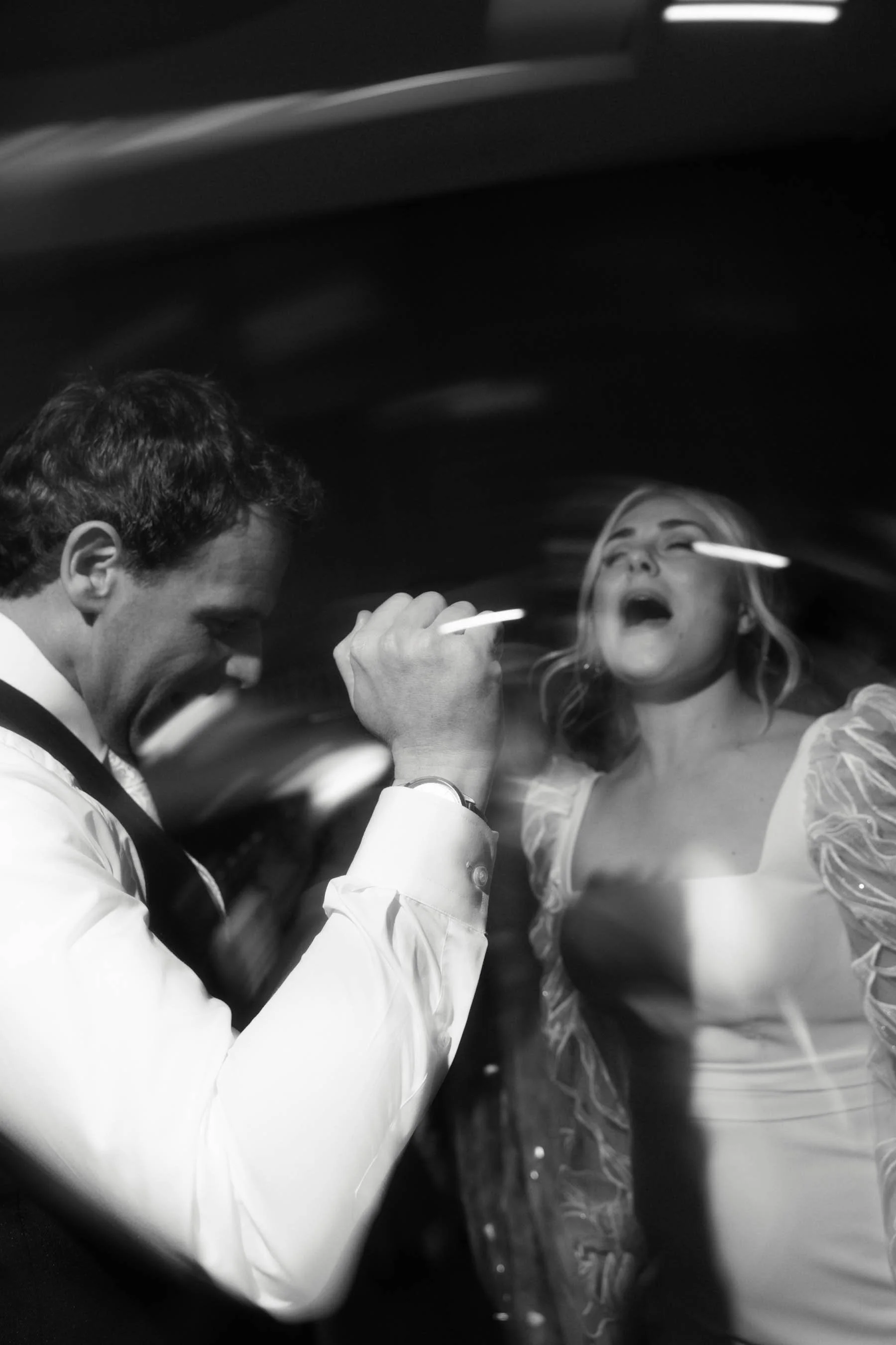 A man in a white shirt and a woman in a patterned dress are having their faces made up or painted by a professional makeup artist with a brush, inside a vehicle or a confined space.