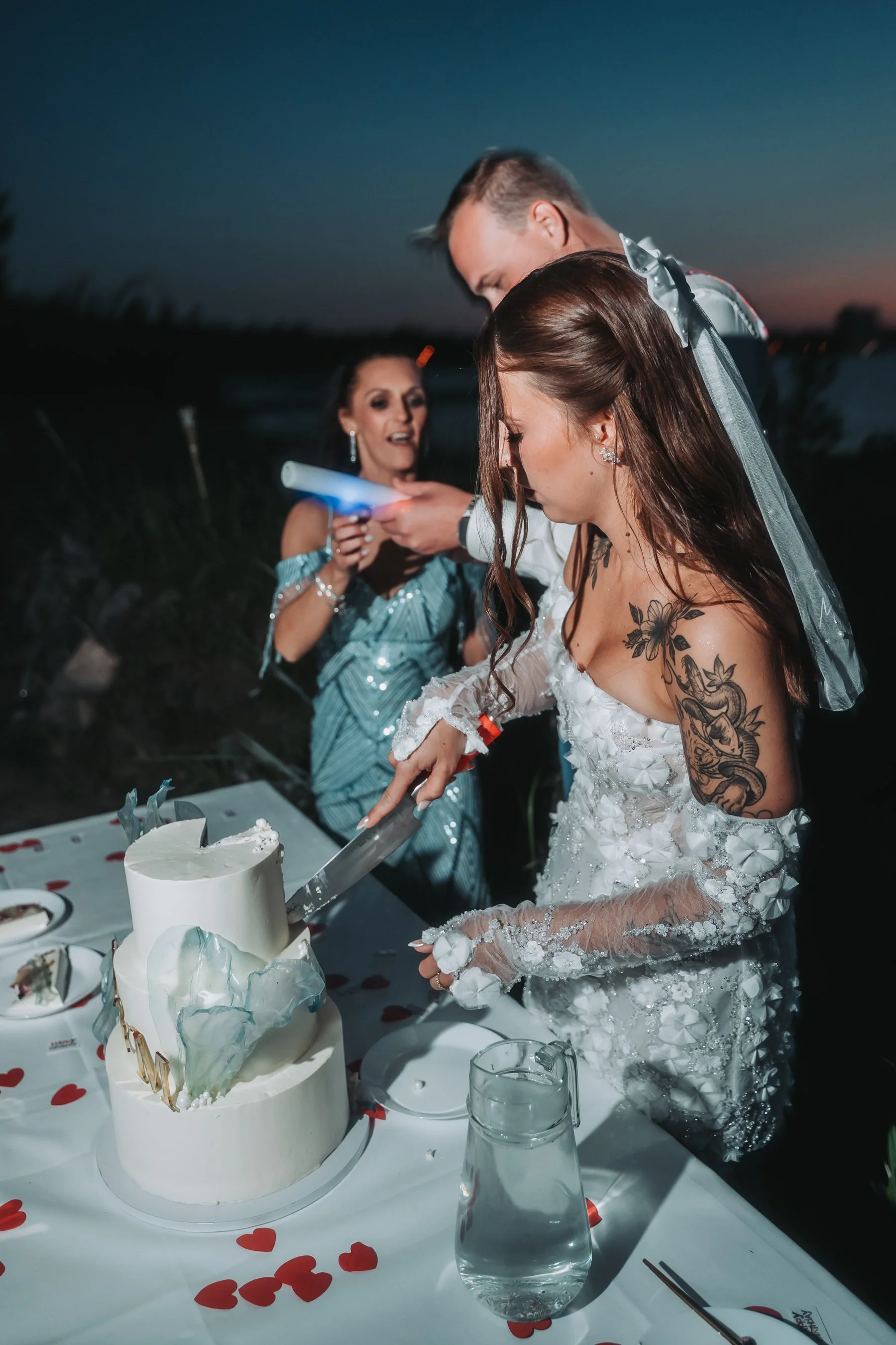 A bride with tattoos on her arm cuts a wedding cake with a large knife, while a woman in a blue dress looks on, during a celebration at sunset.