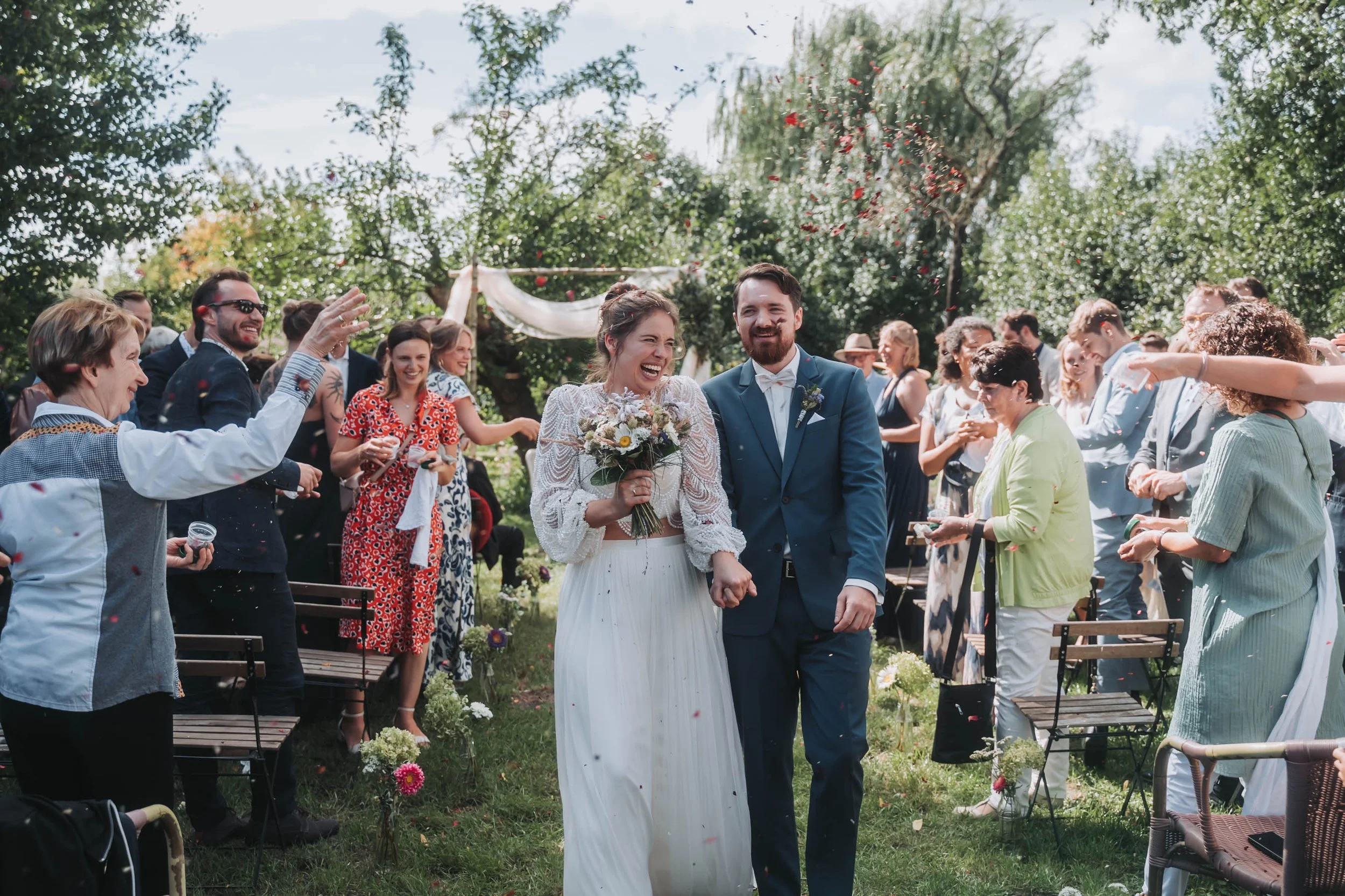 A bride and groom walking hand in hand through a wedding celebration outdoors, surrounded by friends and family throwing flower petals.