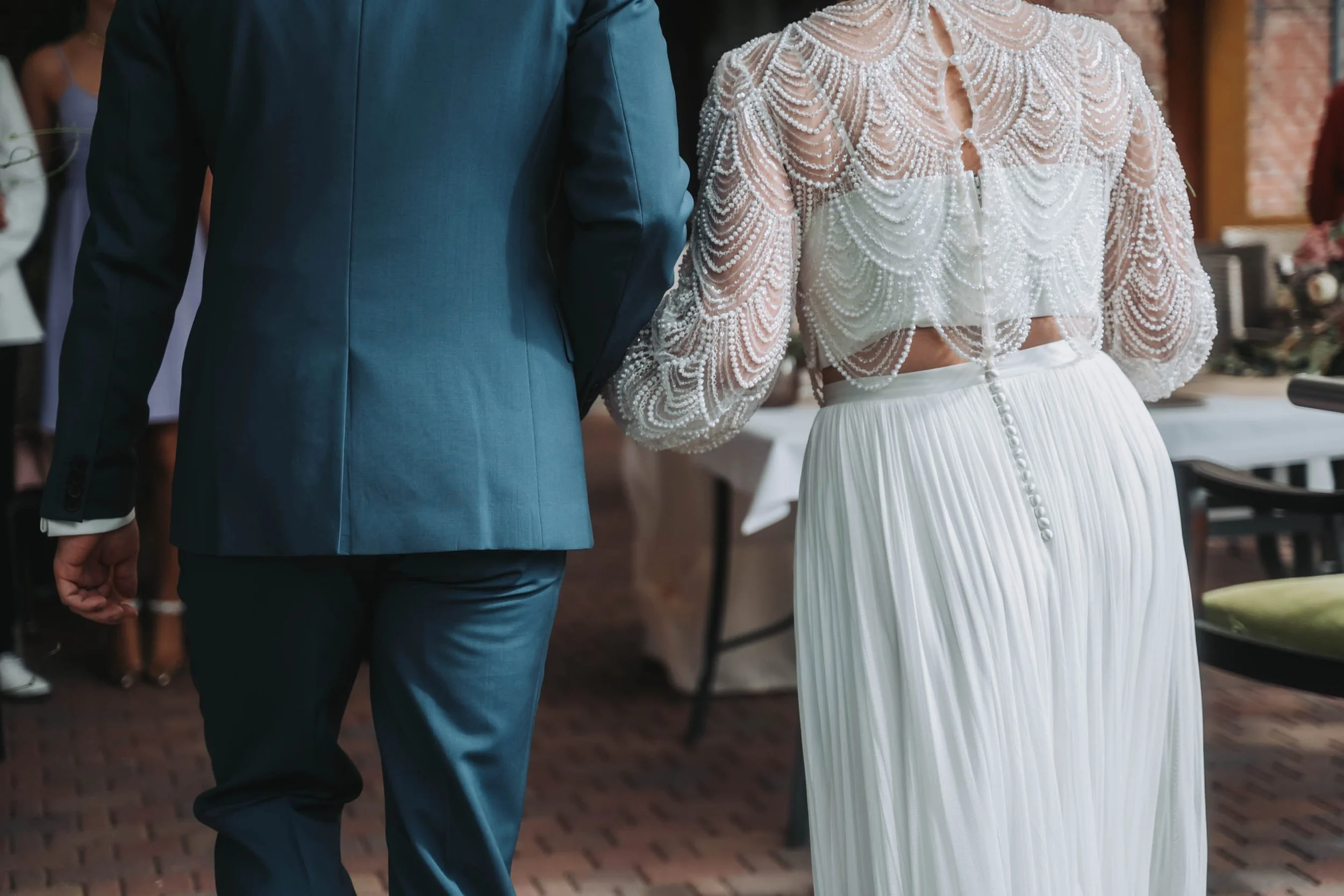A bride and groom walking holding hands at their wedding reception. The bride is wearing a beaded white dress with long sleeves and pleated skirt, and the groom is in a blue suit. The background shows a gathering of guests and a decorated indoor venu