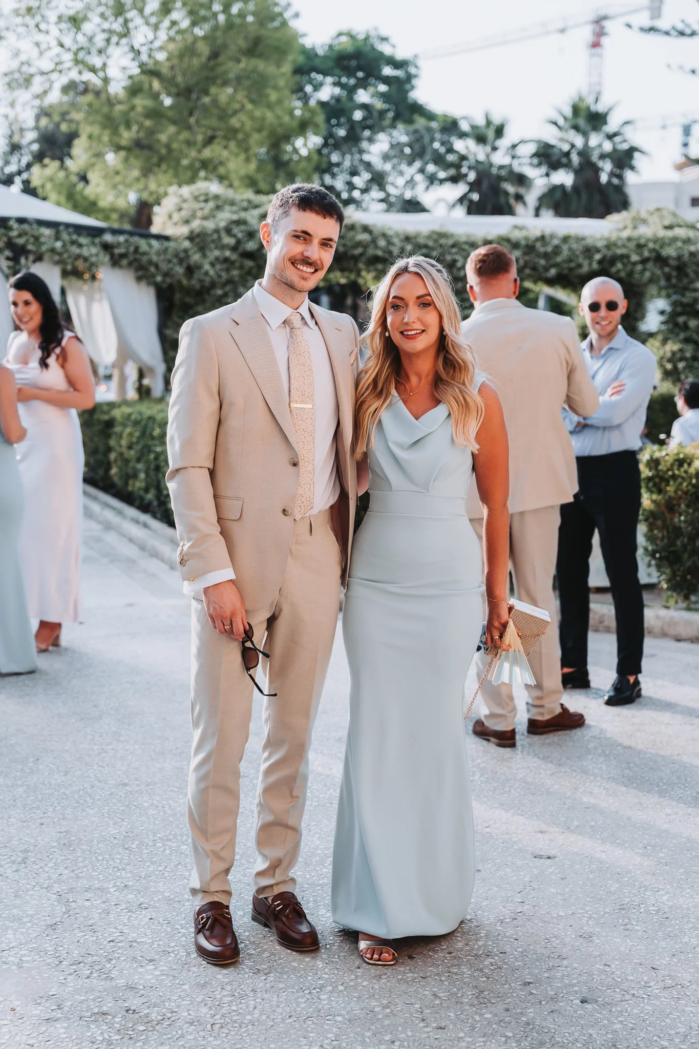 A smiling couple dressed in formal attire at an outdoor event, with others in the background. The man is wearing a beige suit with a white shirt and tie, holding sunglasses, while the woman is in a light blue dress holding small gift bags.