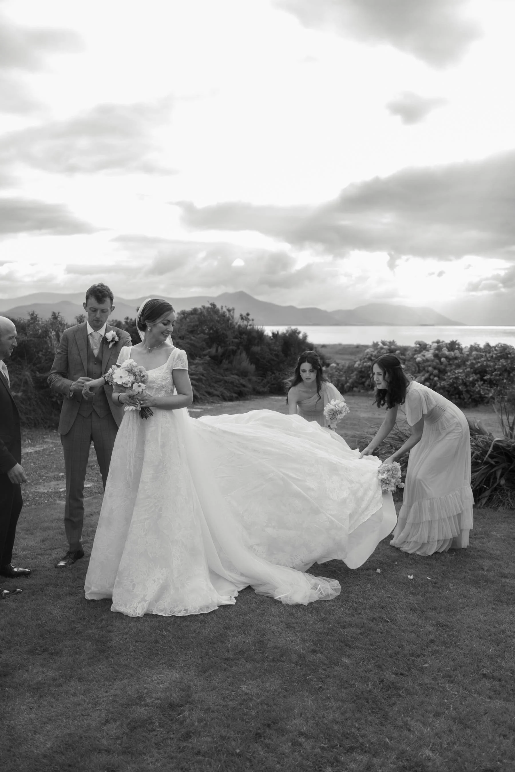 A group of people, including a bride in a wedding dress holding a bouquet and a groom in a suit, are outdoors with mountains and a lake in the background, possibly preparing for or during a wedding ceremony.