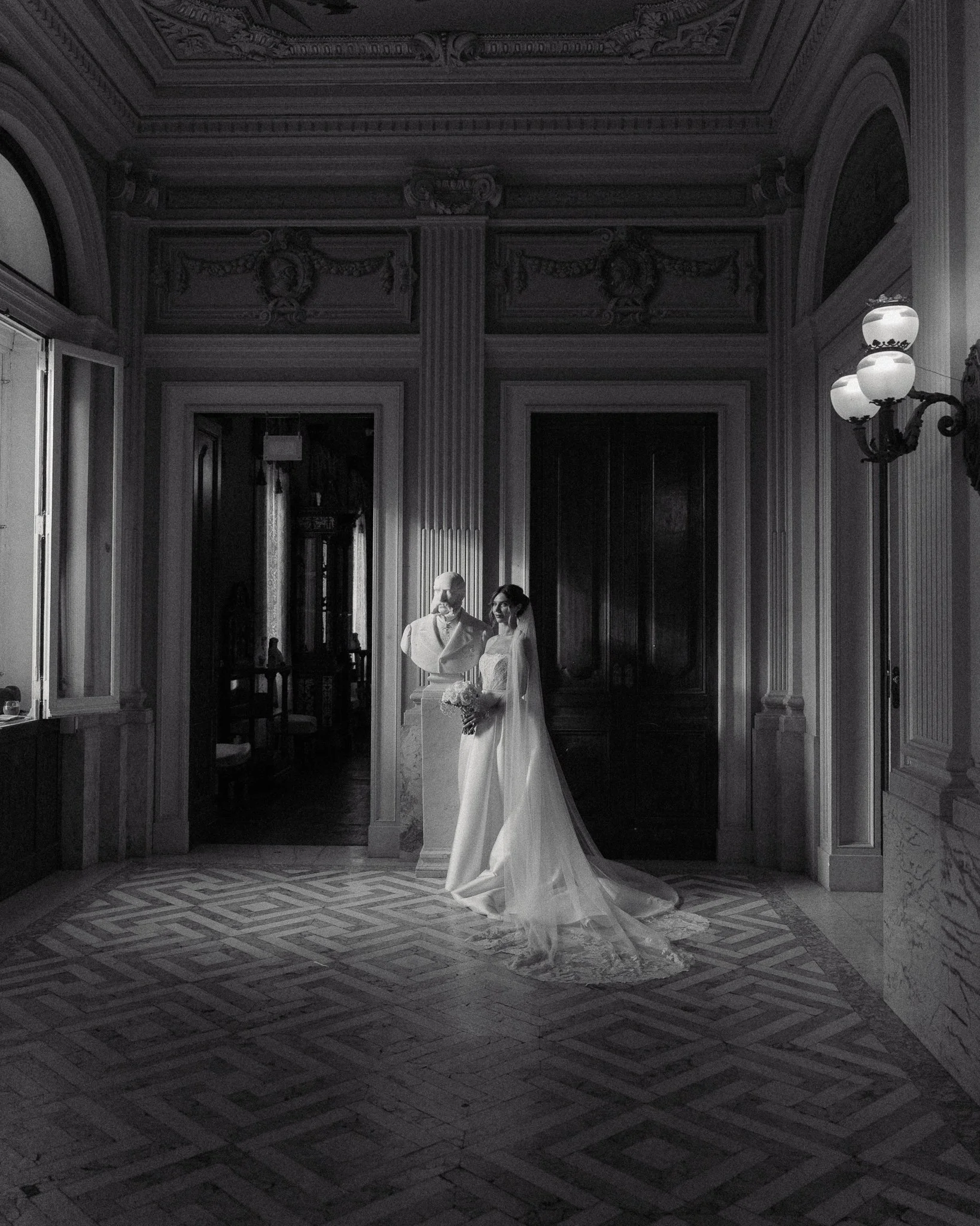 A bride and groom stand together inside an ornate, classical-style room with intricate wall and ceiling decorations. The bride wears a white wedding gown with a veil, while the groom wears a light-colored suit. They are positioned near a bust sculptu