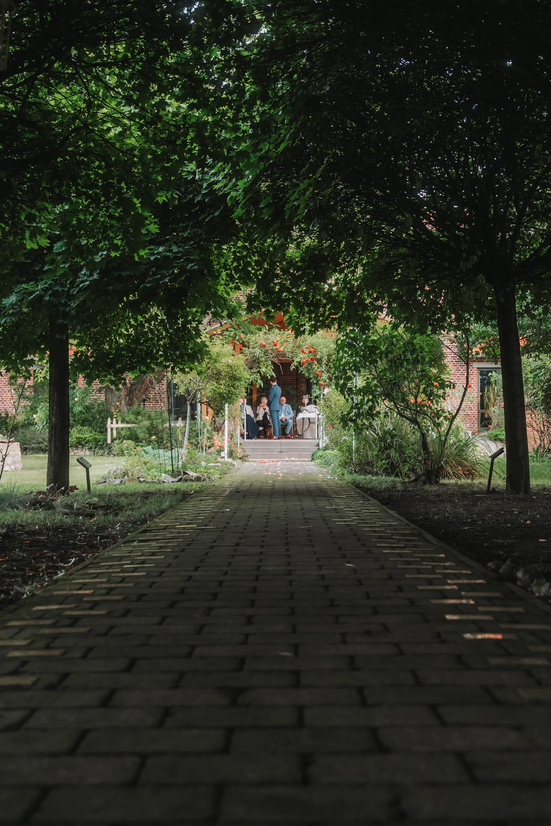 A brick house with a porch and a group of people sitting and standing on the steps, seen through a gap in the leafy branches of trees overhead, with a brick pathway leading to the house.