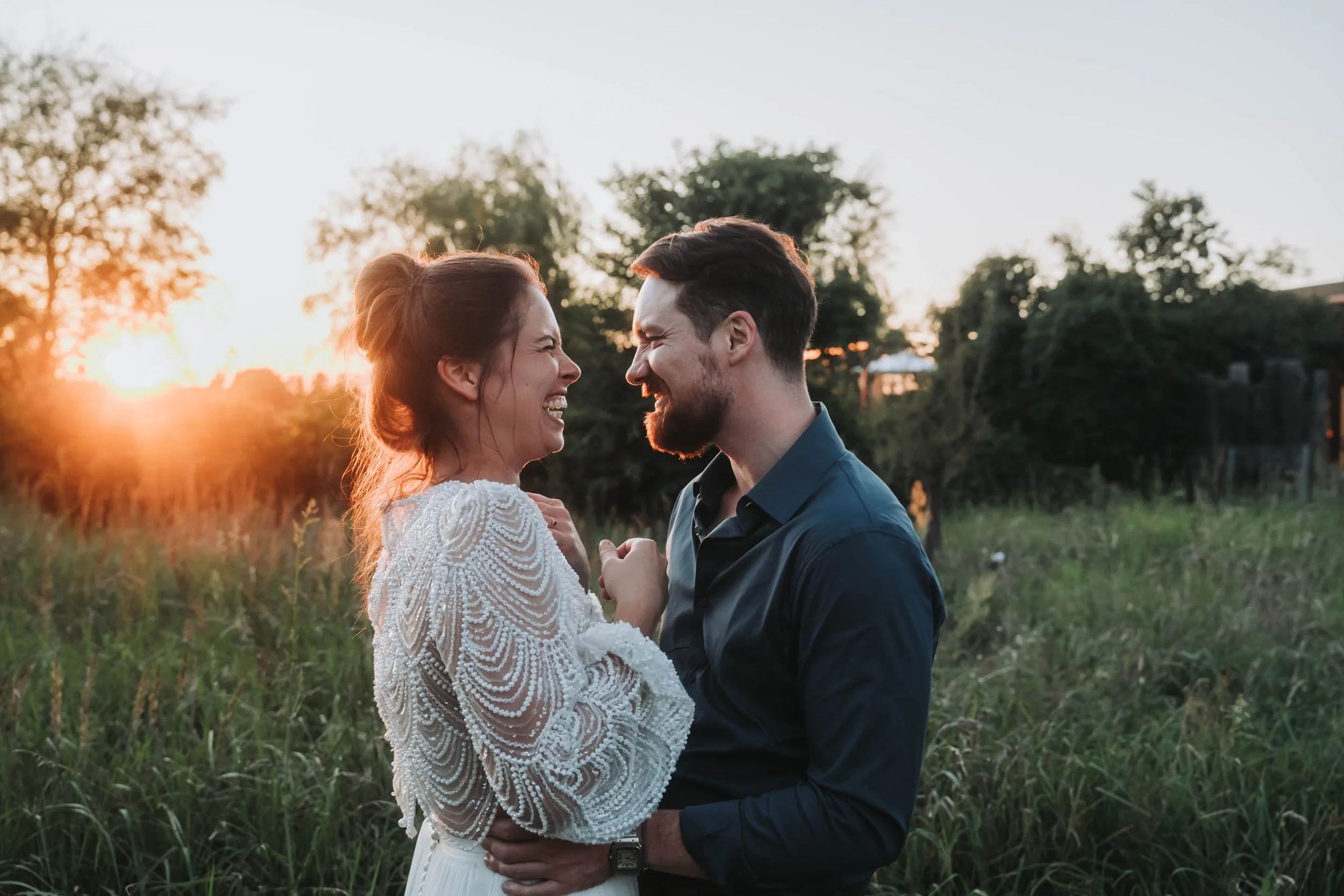 A couple smiling and laughing outdoors at sunset in a grassy field, facing each other closely, with trees in the background.