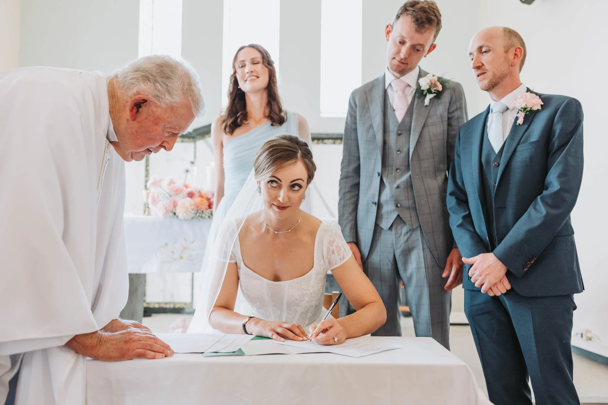 Bride signing marriage documents with family and officiant present.