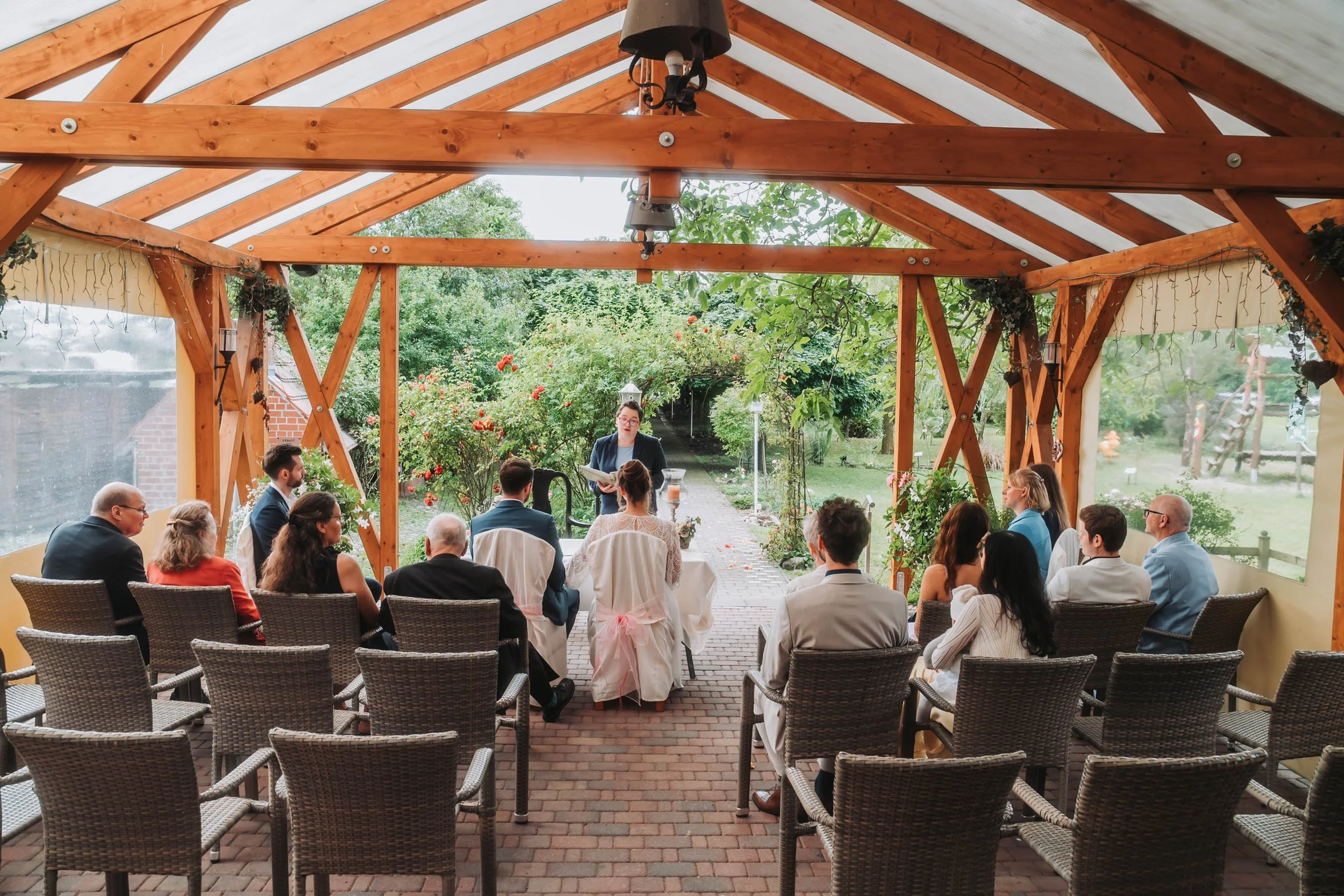 Indoor wedding ceremony with guests seated and a officiant standing in front, under a wooden pergola with transparent roof panels, greenery, and garden background.
