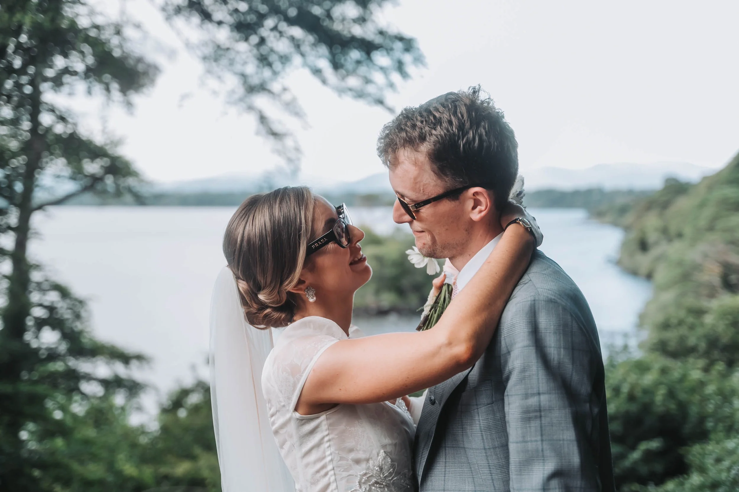 A bride and groom are embracing outdoors, smiling at each other, with a lake and trees in the background.
