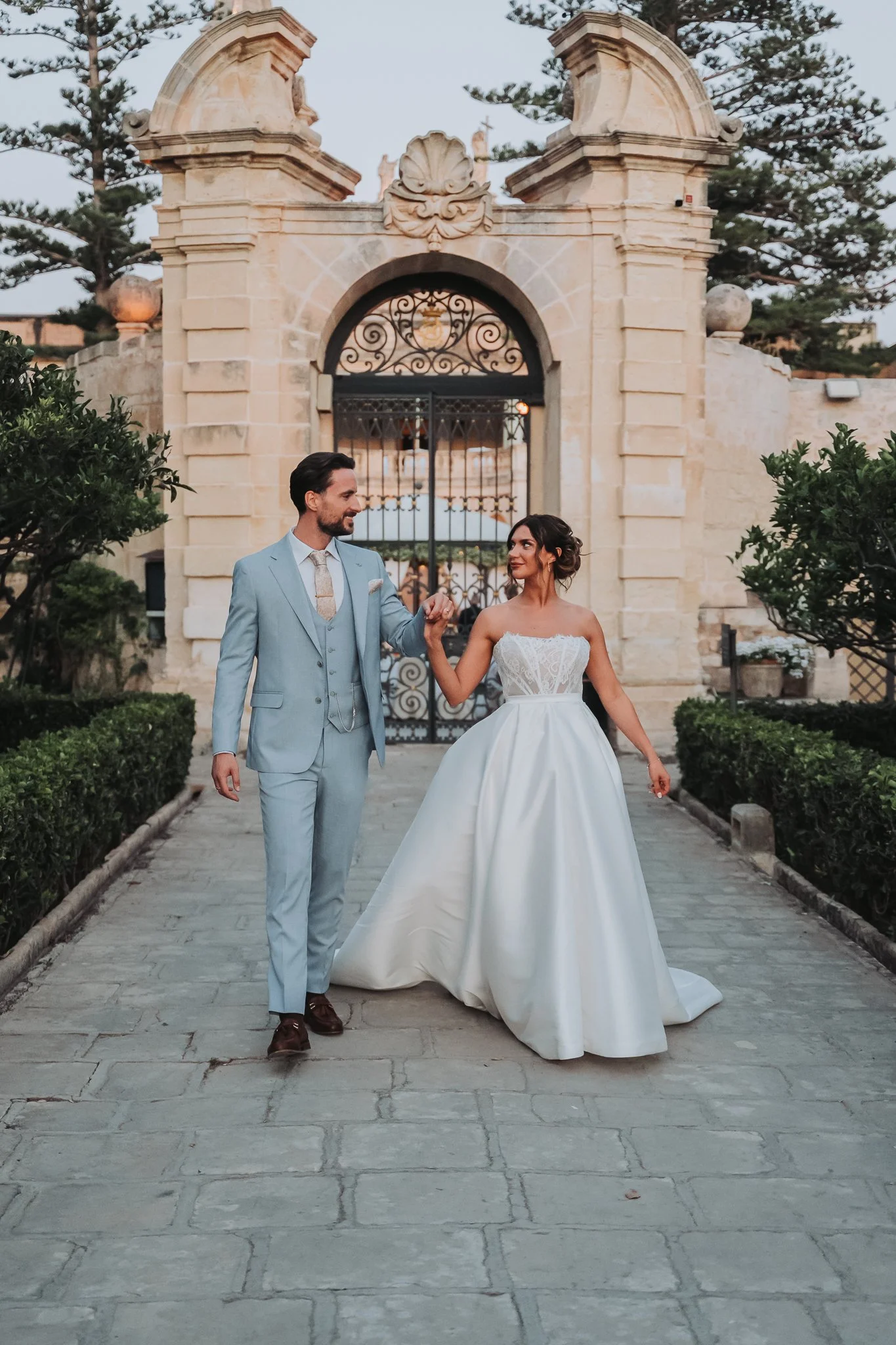 A newly married couple walking outside in front of a historic stone gate. The groom is wearing a light blue suit and the bride is wearing a white wedding dress. They are holding hands and looking at each other. There are trees and greenery around and