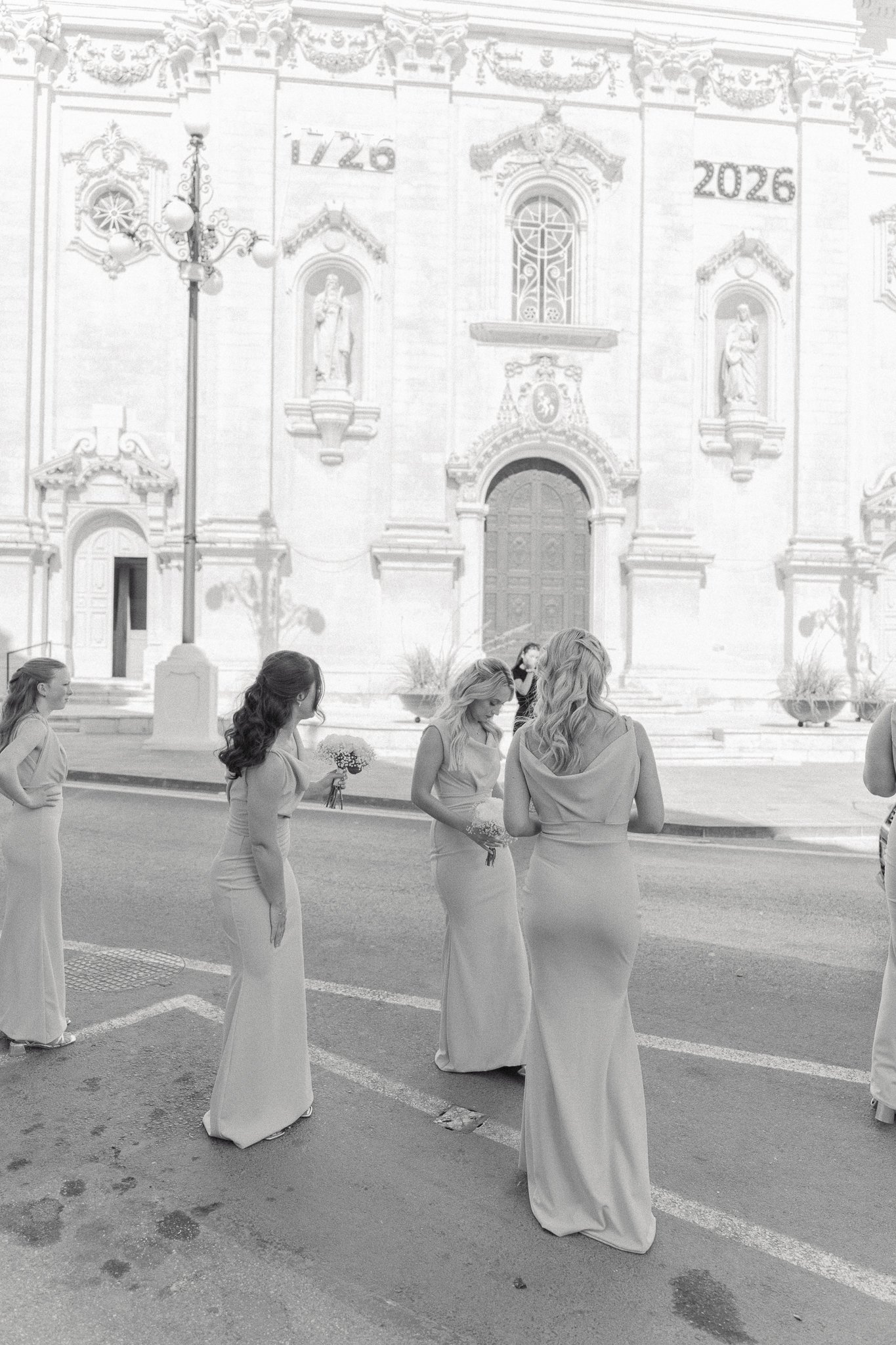 A group of women in elegant dresses standing and conversing outside in front of an ornate historical building with sculptures and the dates 1726 and 2026 displayed on its facade.