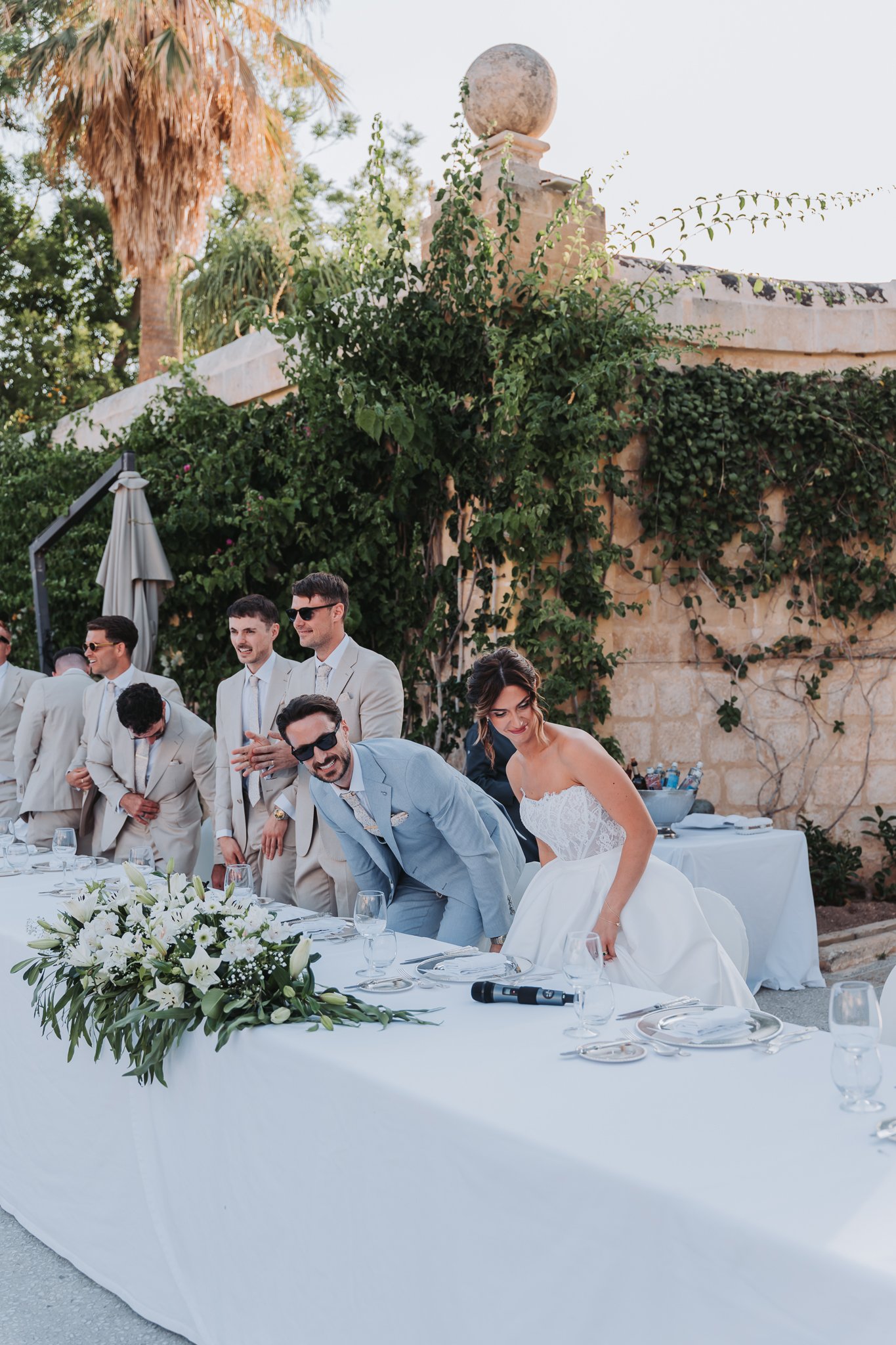 A wedding reception outdoors with a long table decorated with a white tablecloth and a floral centerpiece of white flowers and greenery. Guests dressed in formal attire are standing around the table, some clapping and smiling. Background includes a s