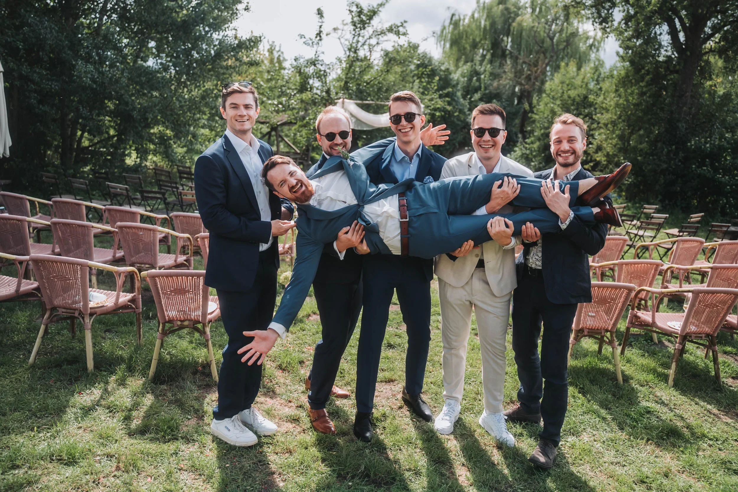 Group of six men outdoors, five standing and one lying across their arms, smiling and laughing.
