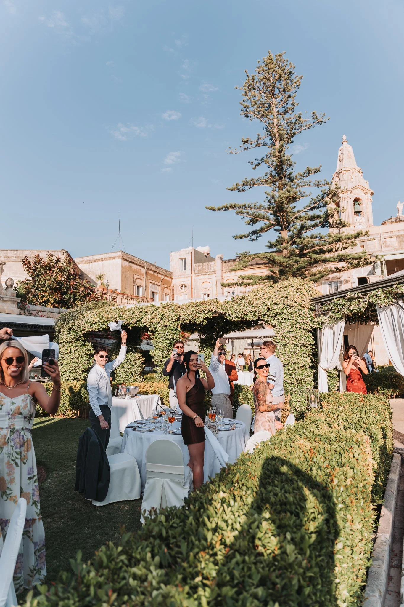 People celebrating at a wedding reception outdoors in a garden, with tables set for dining, surrounded by greenery, and historic buildings and a tall tree in the background.