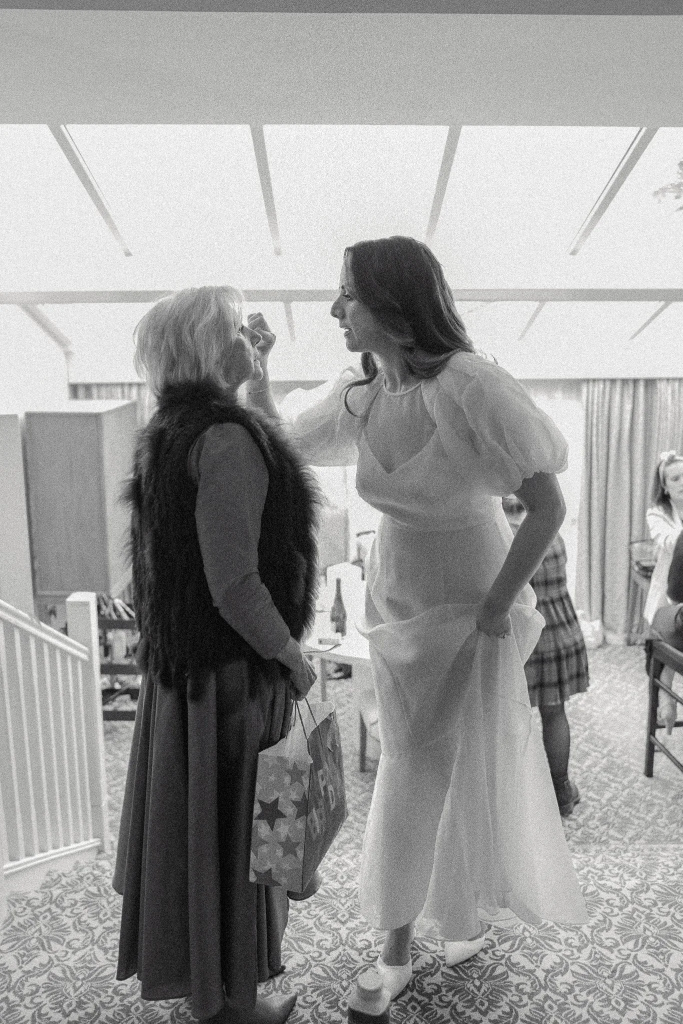 Black and white photo of two women talking inside a room. One woman appears to be holding makeup or skincare tools, and the other is wearing a dress and high platform shoes. Other people are visible in the background.