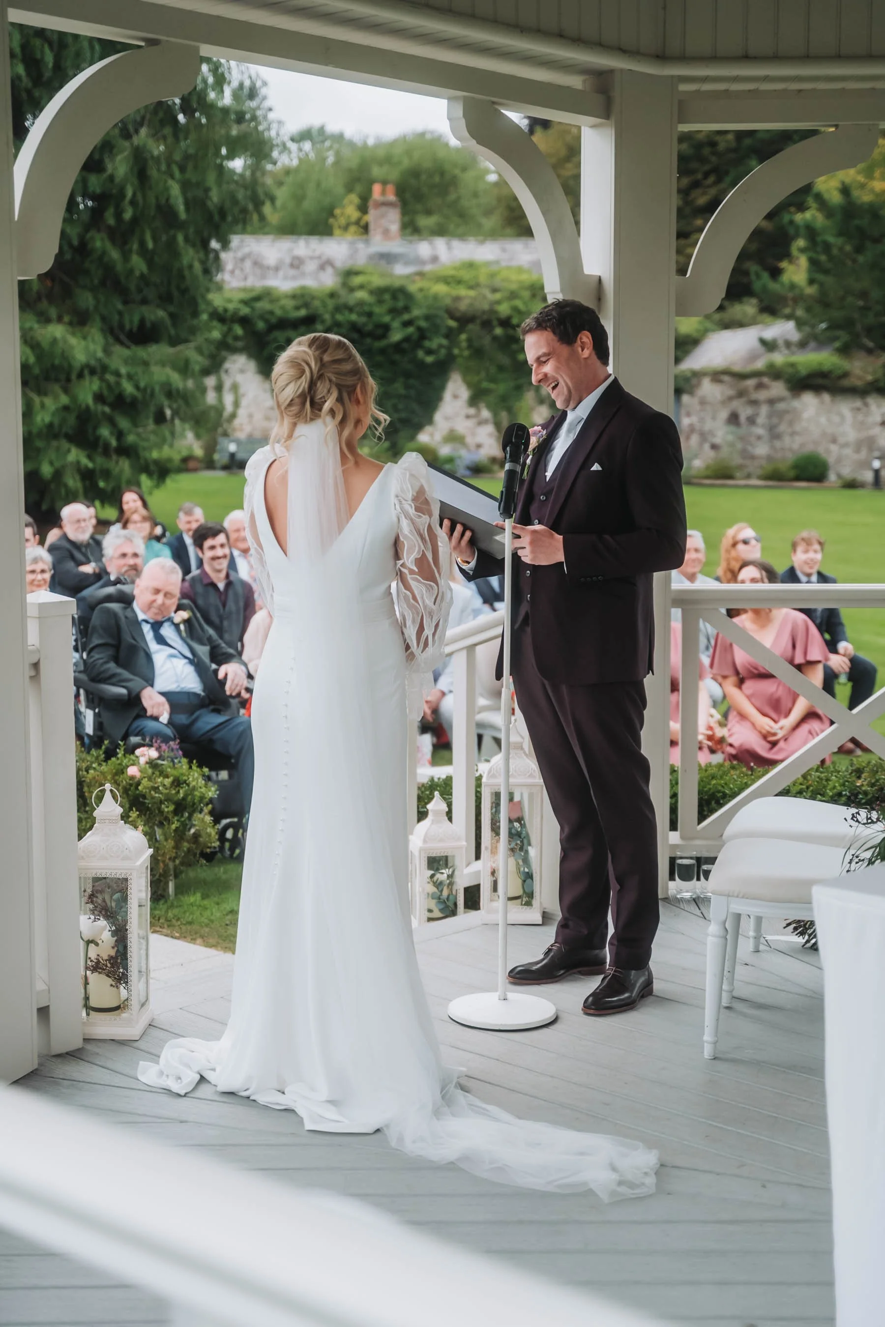 A man and woman exchange vows during a wedding ceremony outdoors on a gazebo. The woman wears a white dress with lace sleeves and a veil, while the man wears a black tuxedo. Guests are seated behind them, watching the ceremony.