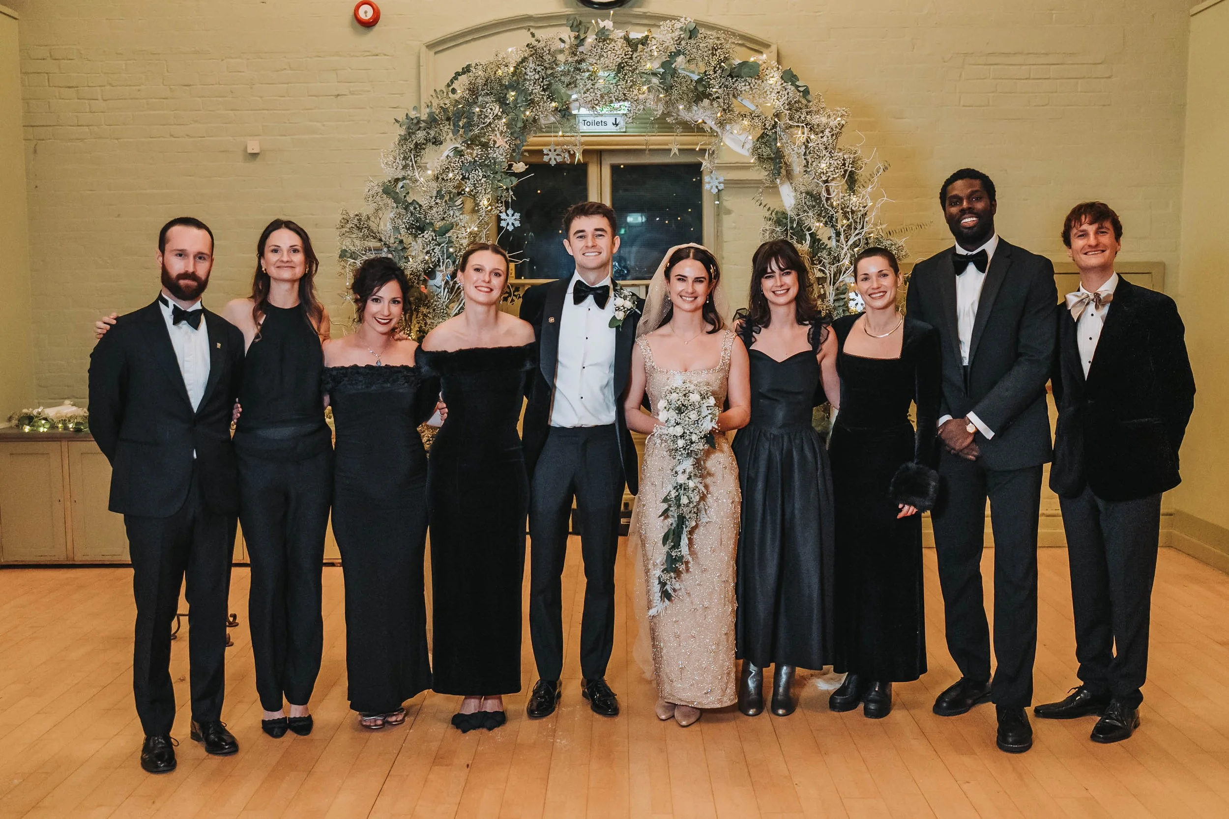 A group of eleven people dressed in formal attire, smiling and posed for a photo in front of a decorated arch at a celebration or event, likely a wedding.
