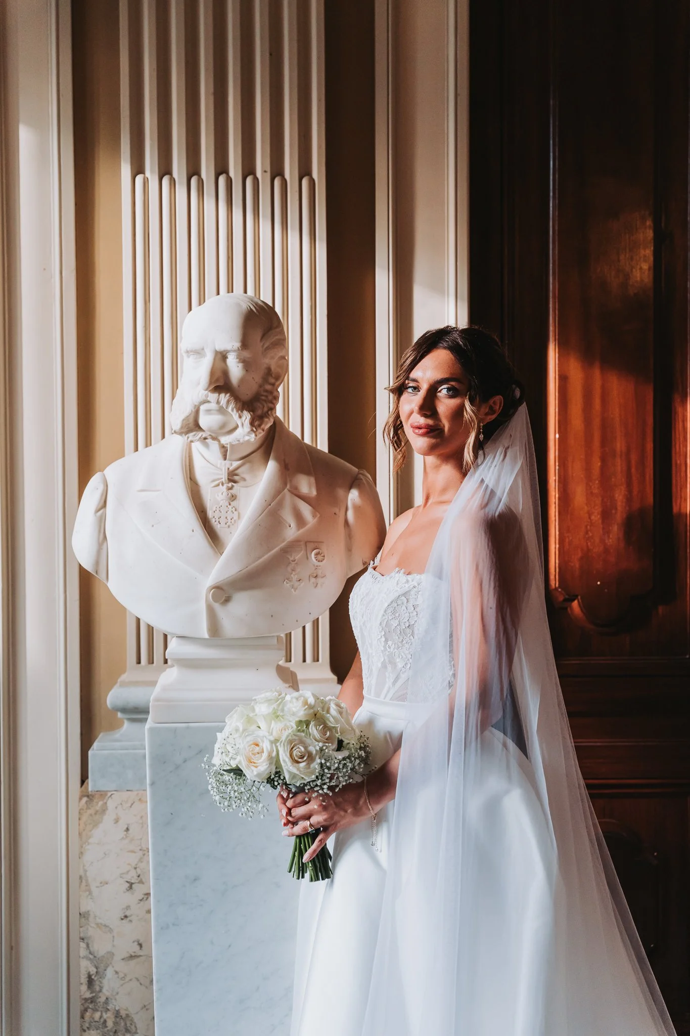 A bride in a white wedding dress holding a bouquet of white roses standing next to a white bust sculpture of a man with a beard, featuring a tuxedo and bowtie, in an elegantly decorated room with wood paneling and marble accents.