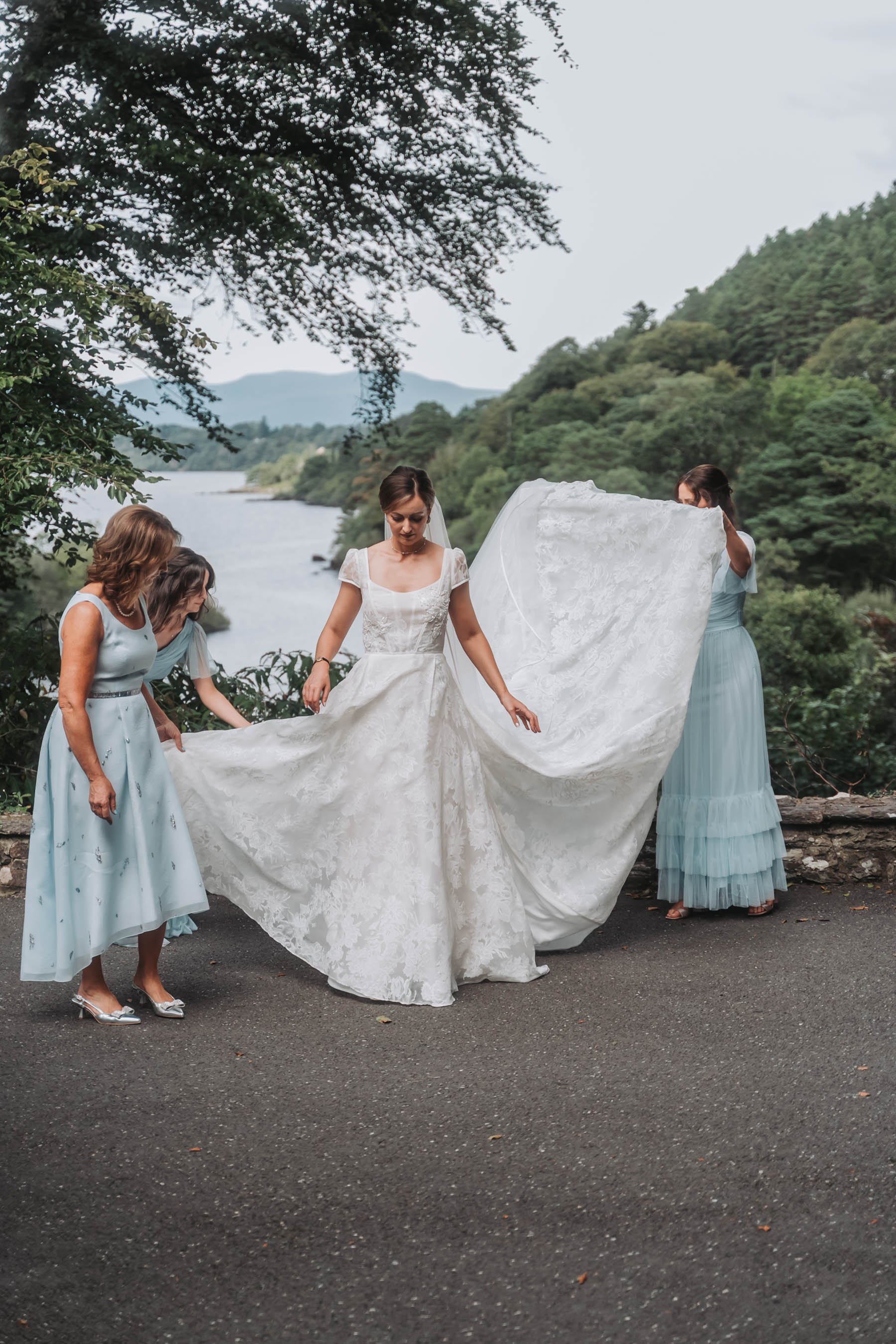 A bride in a white wedding dress with lace details stands outdoors near a river and forested hills, surrounded by four women dressed in pastel blue dresses, helping her with her gown. One woman is holding a large section of the gown, while another is