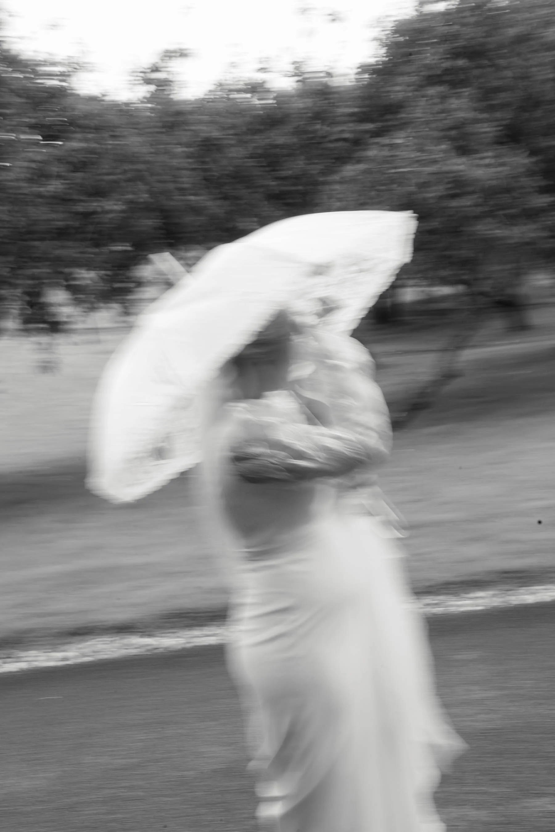 Black and white photo of a person walking outdoors, carrying or wearing a large umbrella. The background features blurred trees and bushes, suggesting motion or movement.