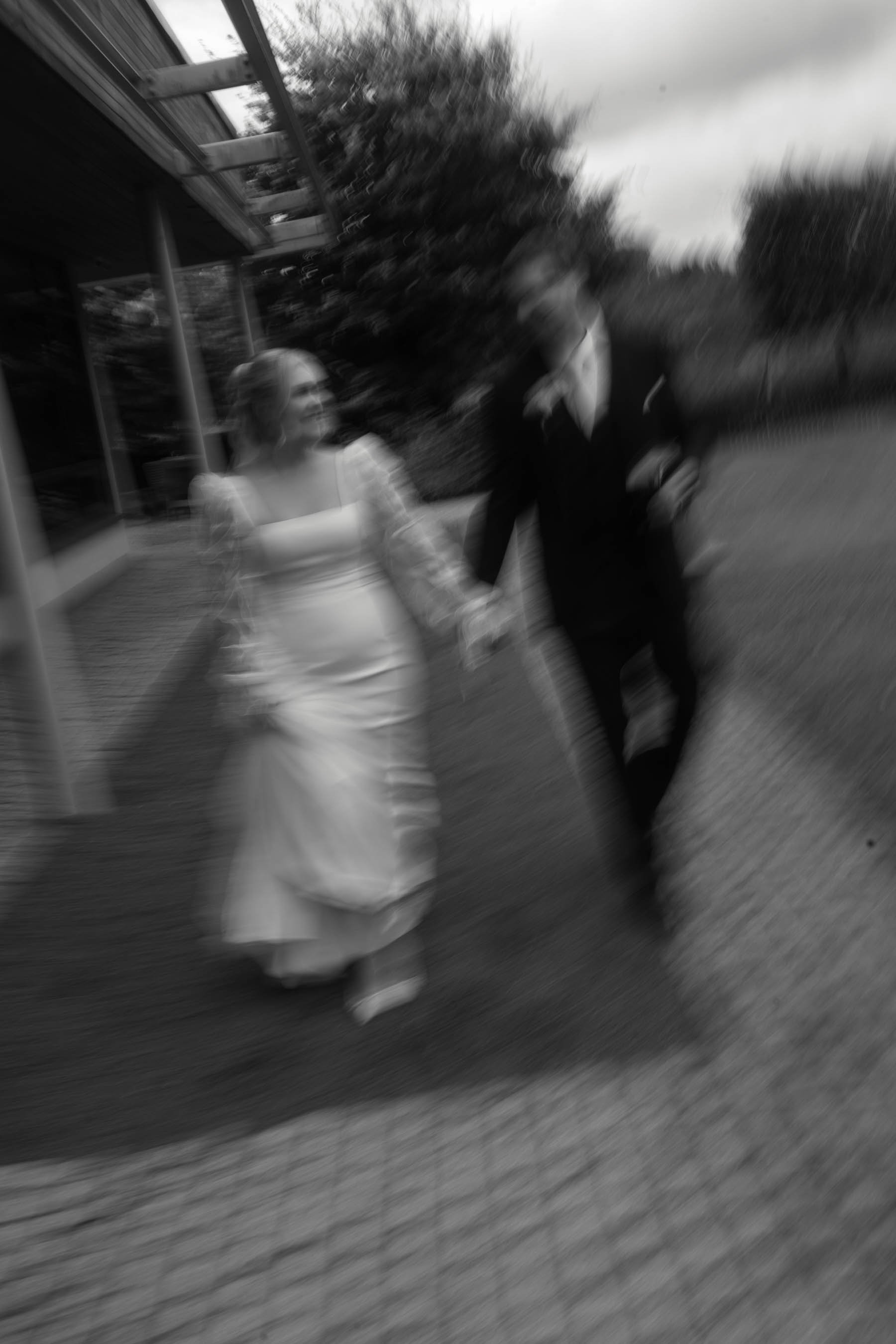 Blurred black-and-white photo of a woman in a white dress and a man in a dark suit walking outside near a building and trees.