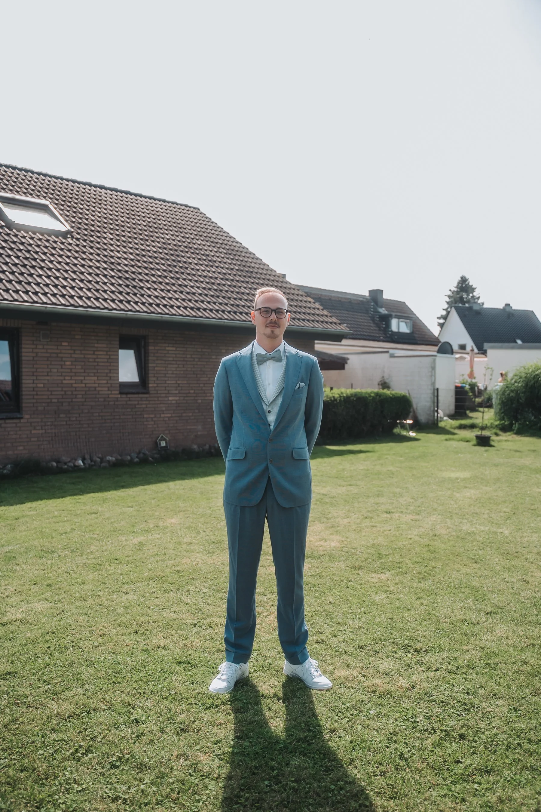 A man in a light blue suit and white sneakers stands on a grassy yard with houses in the background, under a clear sky.