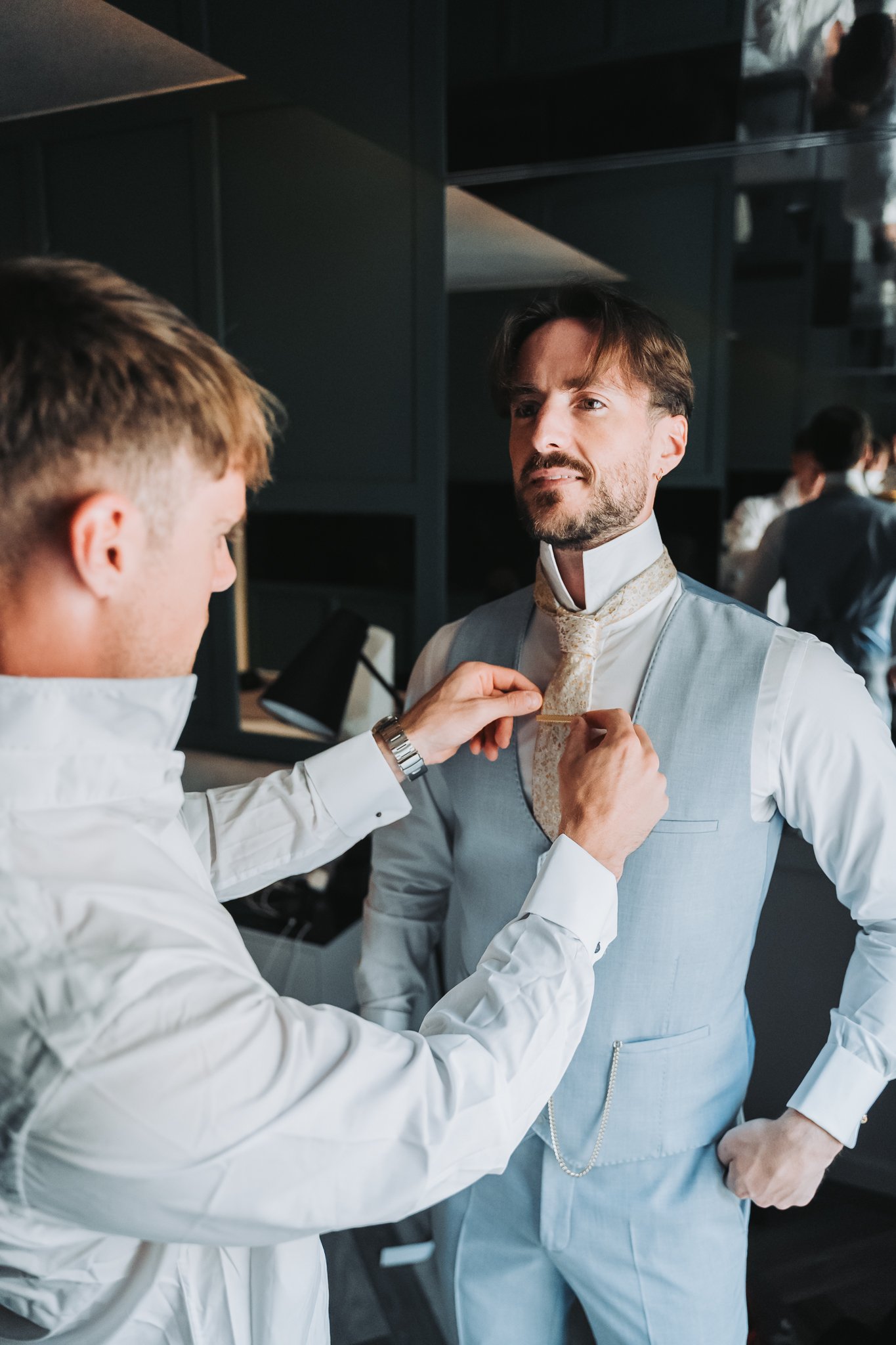 Two men dressed in formal attire, one adjusting the other's tie in an indoor setting with people and furniture in the background.