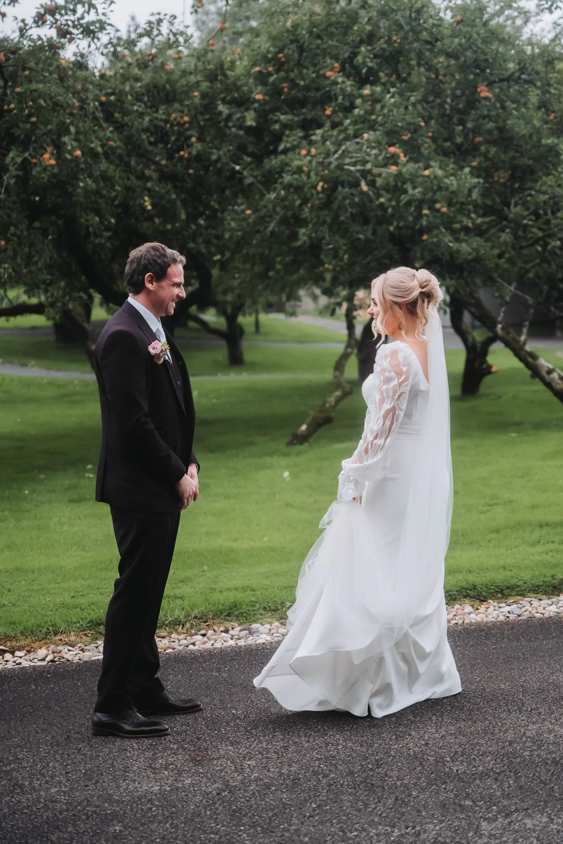 A groom and bride standing on a paved road in a park, facing each other and smiling, with trees and grass in the background. The groom wears a dark suit with a pink boutonniere, and the bride wears a white wedding dress with long lace sleeves.