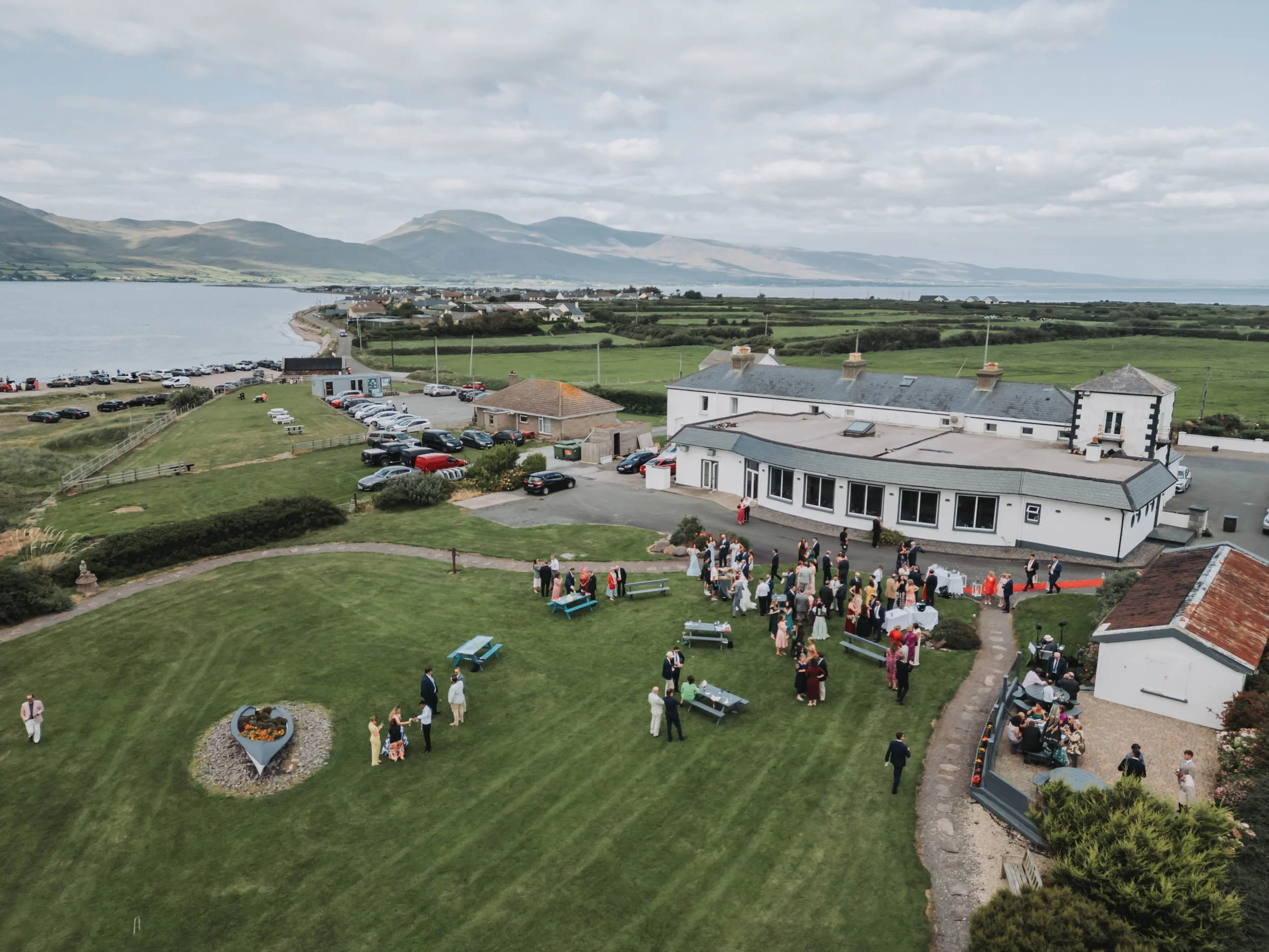 An outdoor wedding reception area with guests gathered on a grassy lawn, some seated at tables and others standing, near a white building with a flat roof. There is a view of a lake, green fields, and mountains in the distance under a cloudy sky.